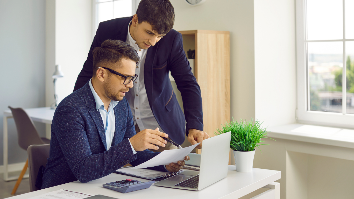Two men in suits reviewing documents near a laptop and calculator in an office.