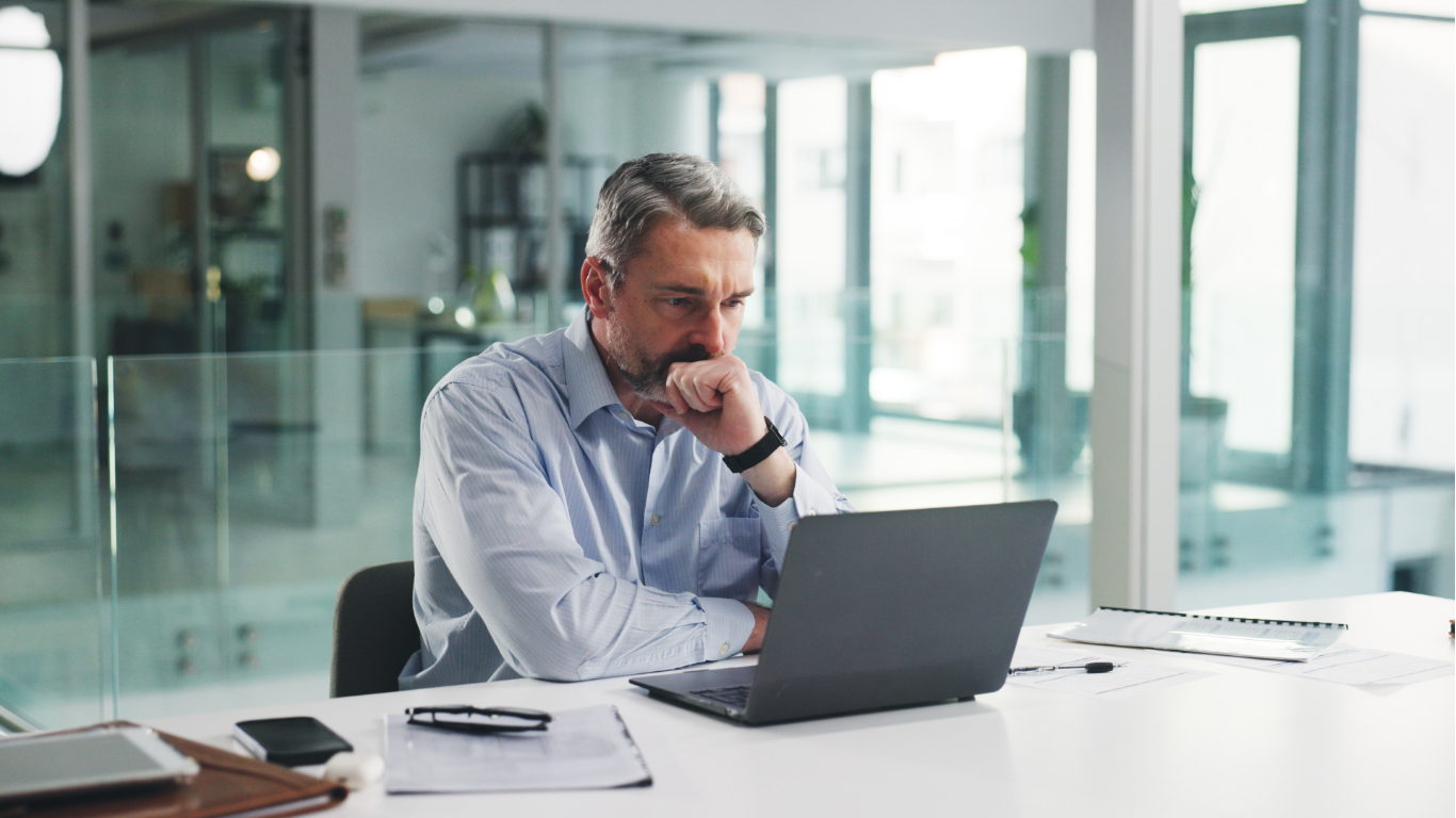 Man at desk, looking at laptop. He’s wearing a blue shirt and touching his chin. Office setting, focused.