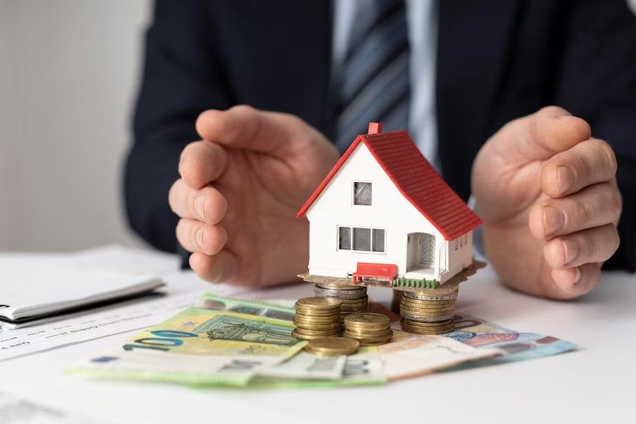 A man is holding a model house on top of stacks of money.