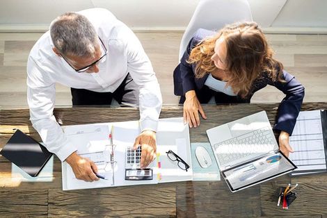 A man and a woman are sitting at a desk looking at papers and a calculator.