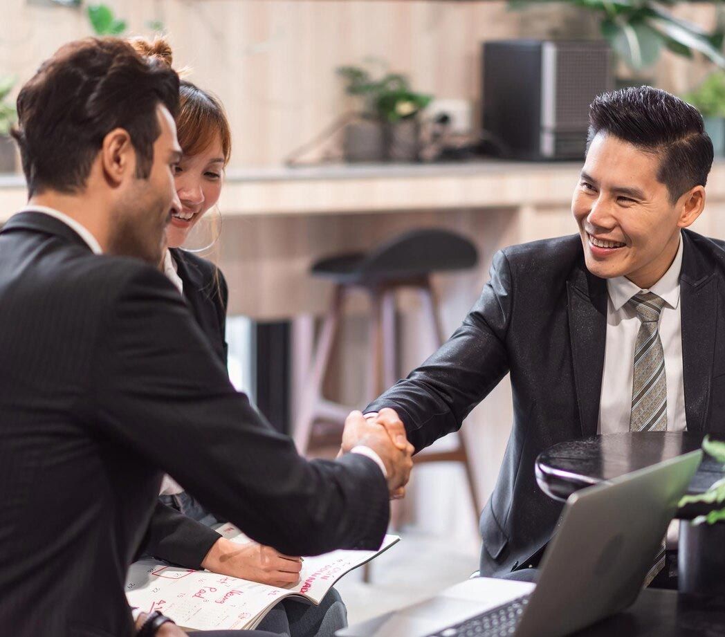 Two men in suits shake hands, smiling, in a business meeting. A woman sits nearby.