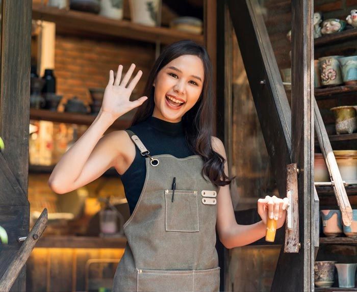 A woman in an apron is standing in front of a glass door and waving.
