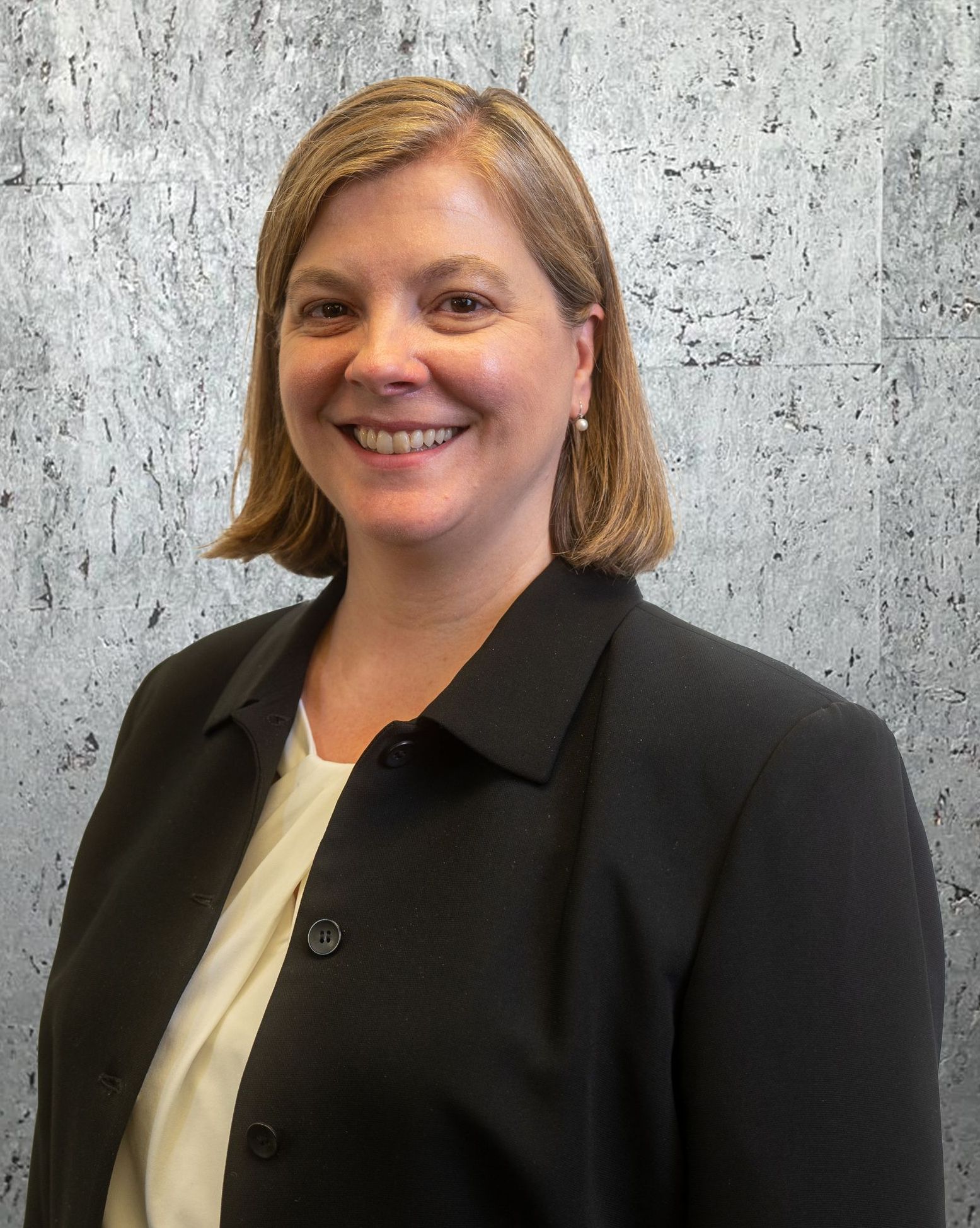 A woman in a black jacket and white shirt is smiling in front of a concrete wall.