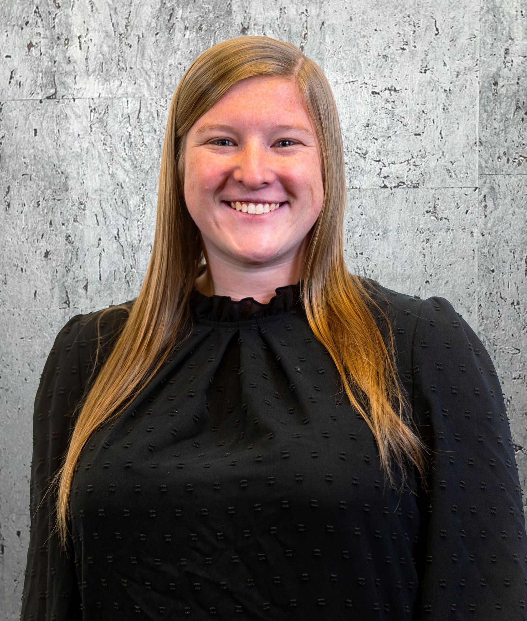 A woman in a black shirt is smiling in front of a concrete wall.