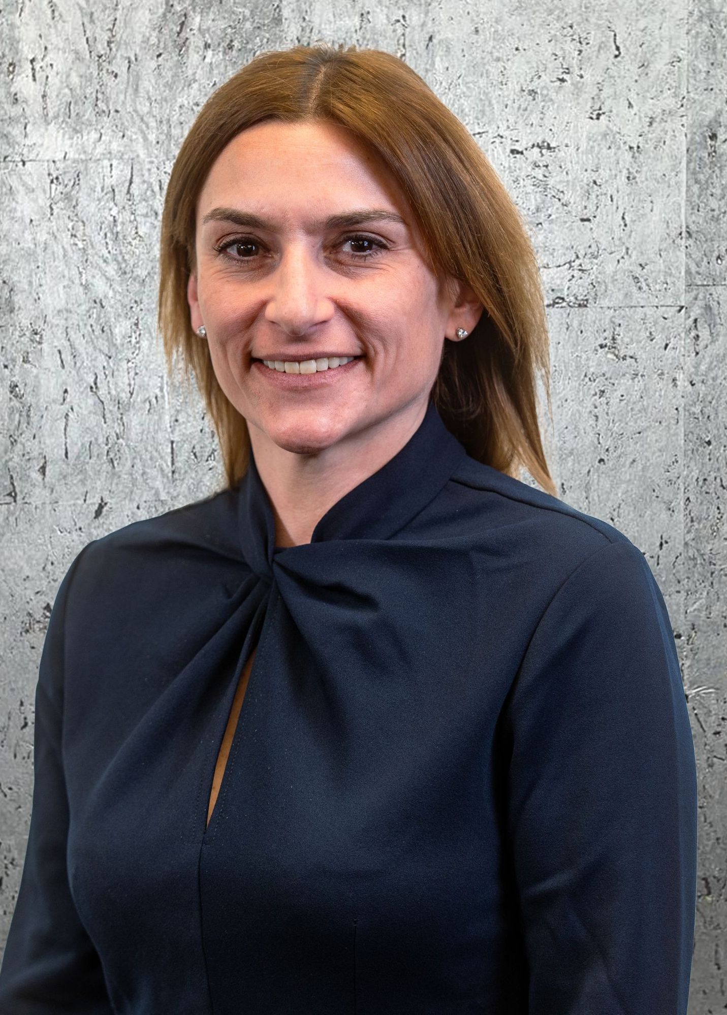 A woman in a blue shirt is smiling in front of a concrete wall.