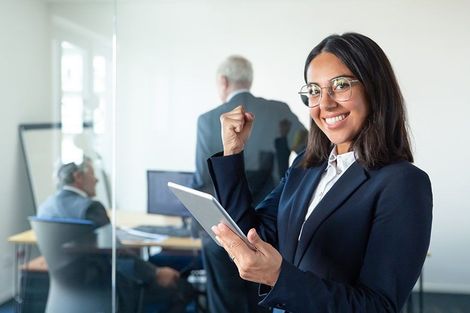 A woman in a suit is holding a tablet and smiling in an office.