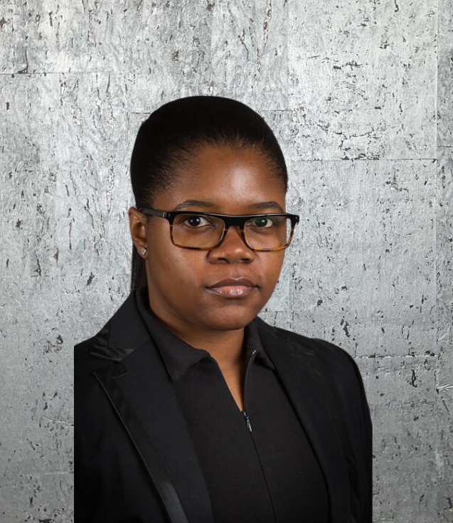 A woman wearing glasses and a black suit is standing in front of a concrete wall.