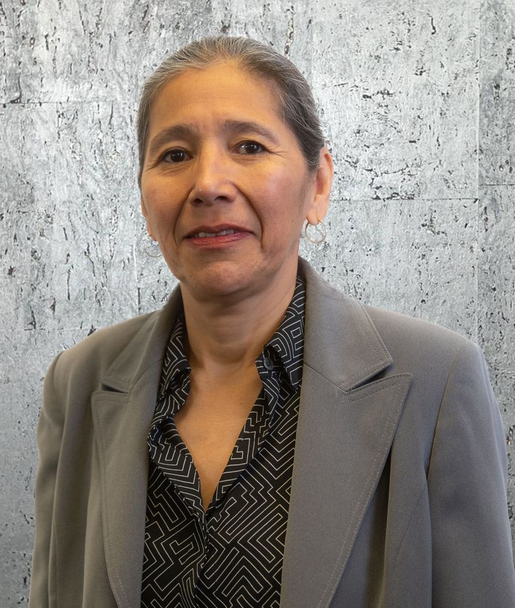 A woman wearing a grey jacket and a black shirt is standing in front of a concrete wall.