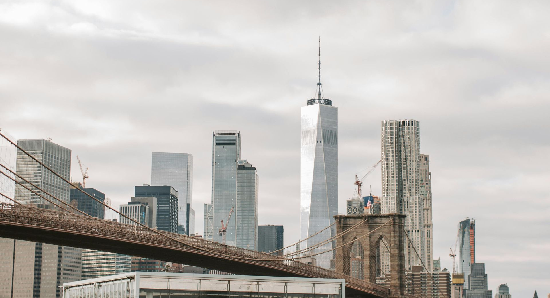 Brooklyn Bridge in foreground, New York City skyline with One World Trade Center against overcast sky.