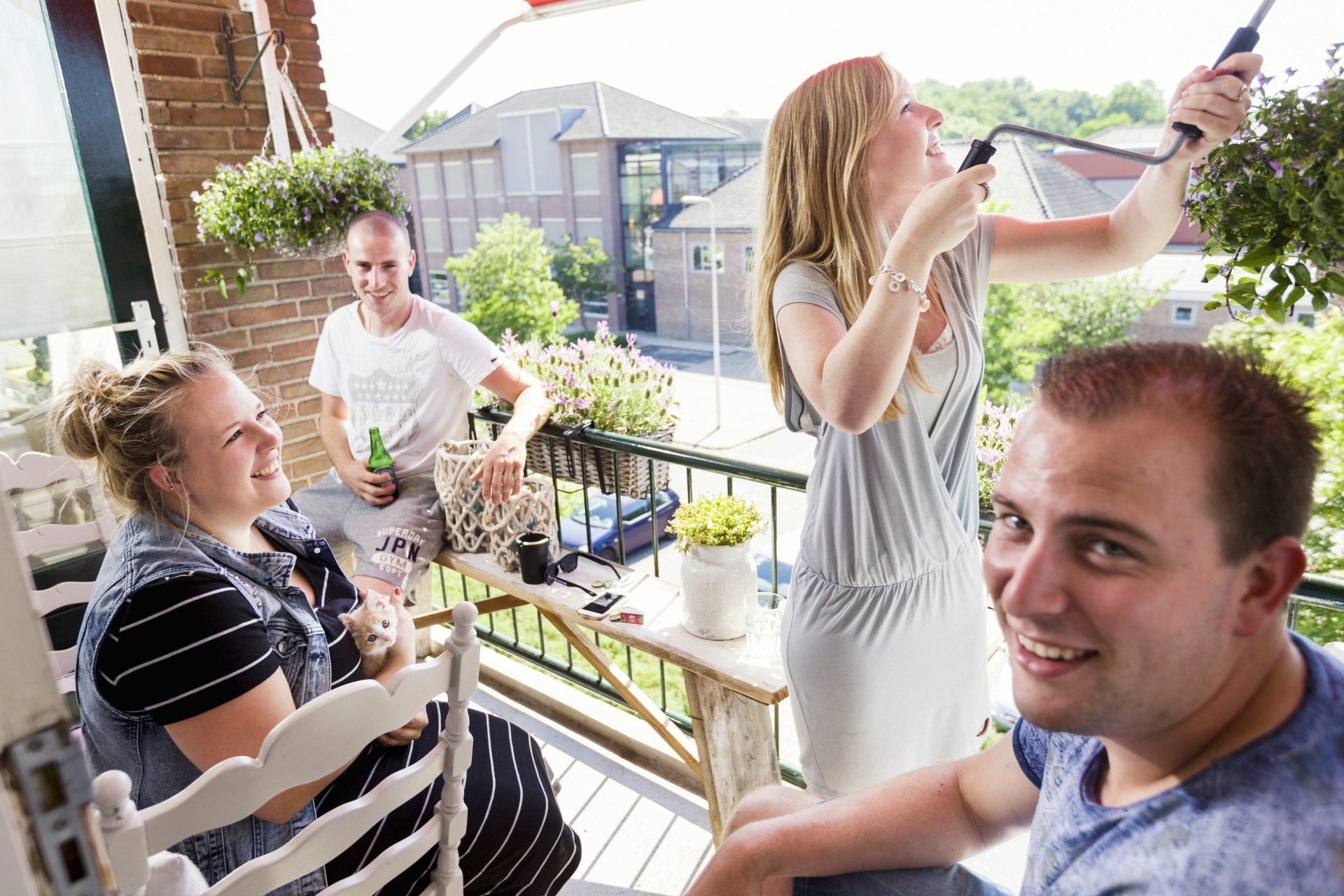 Vier mensen op een balkon op een zomerse dag.  Een vrouw draait het zonnescherm uit.