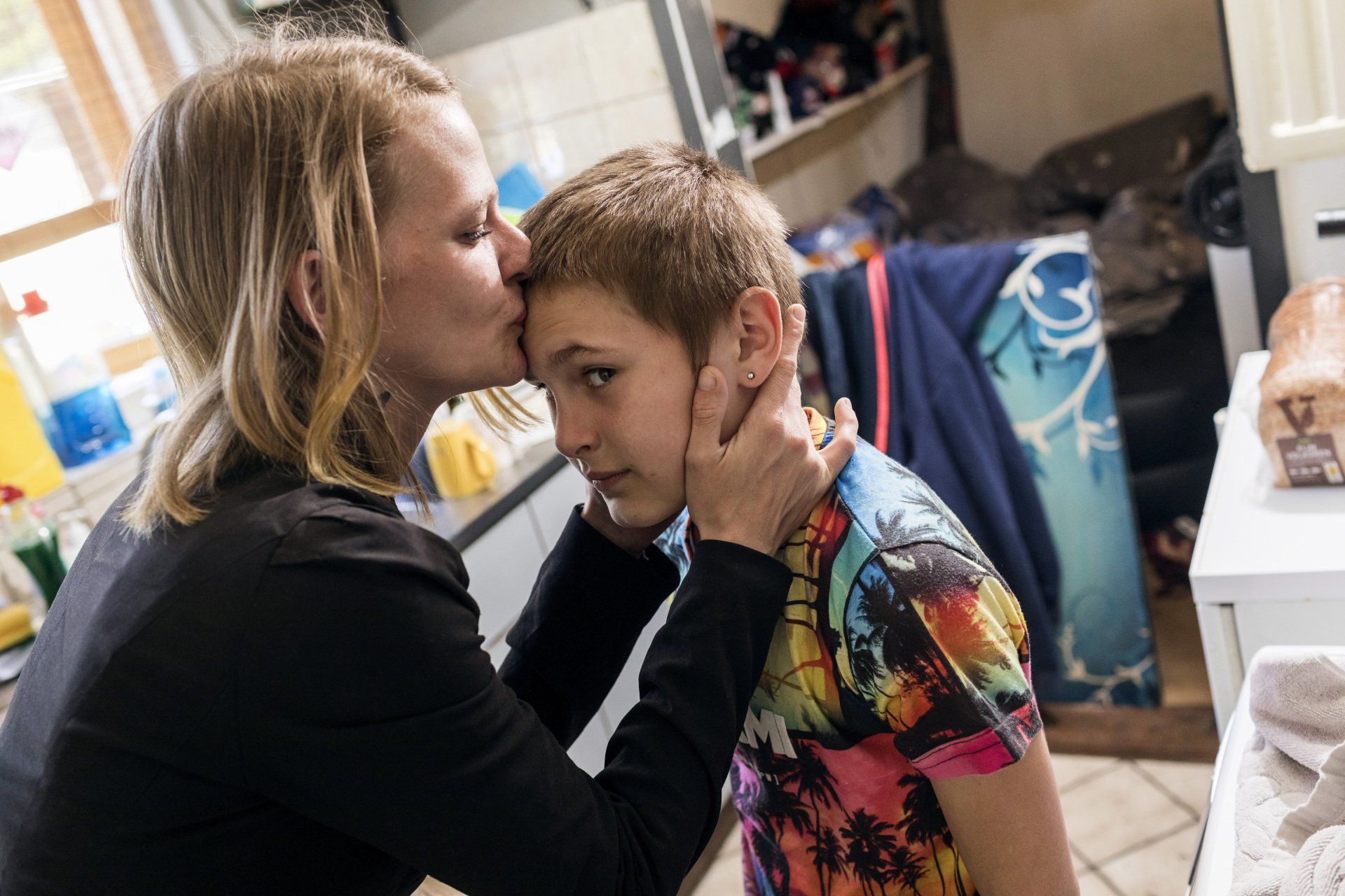 Een vrouw kust een jongen op zijn voorhoofd. Ze houdt zijn gezicht vast. Ze zijn binnen.
