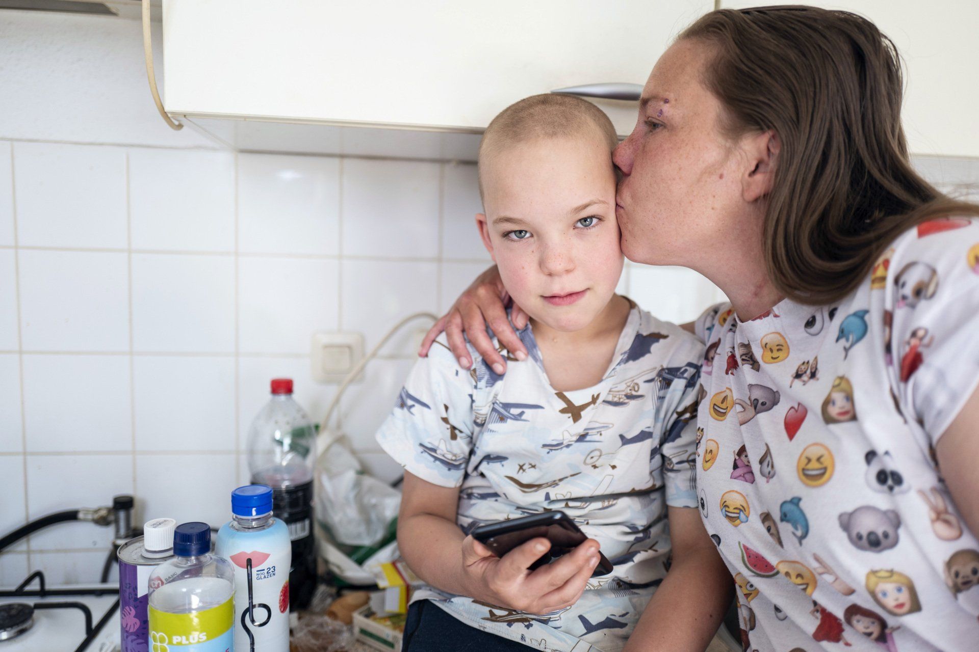 Een vrouw kust een kind op de wang in een keuken. Het kind houdt een telefoon vast. 