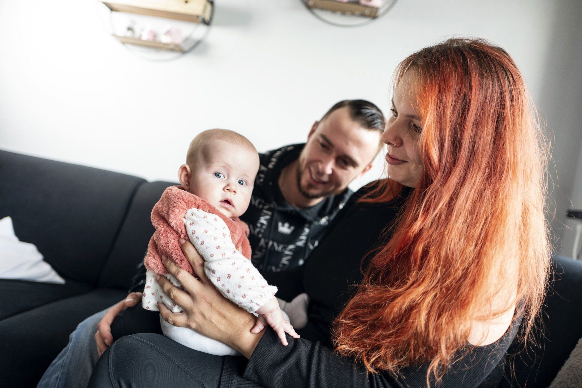Een vrouw met rood haar houdt een baby vast, een man glimlacht achter hen op een bank.