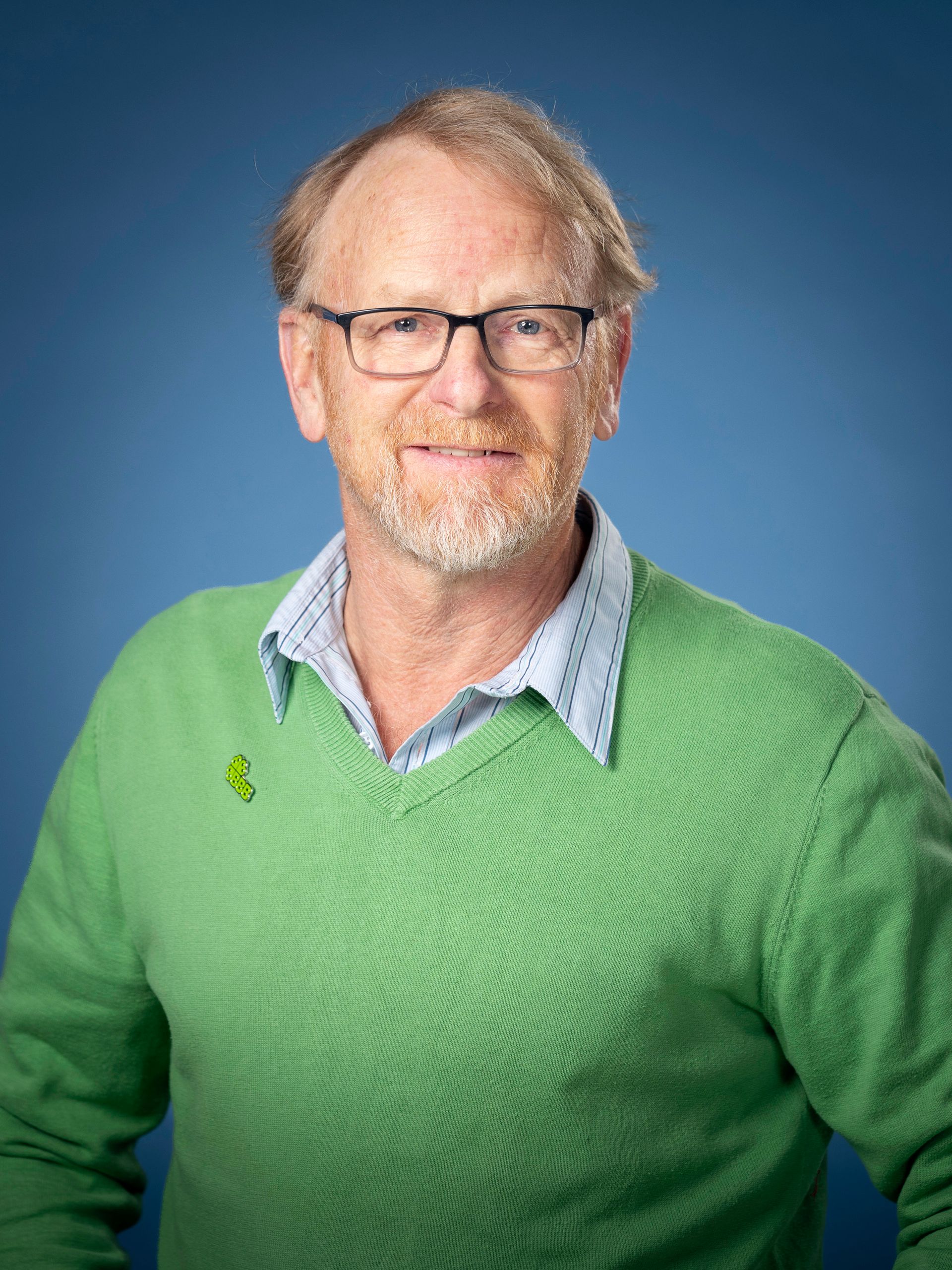 Man met bril, gekleed in een groene trui en een lichtblauw overhemd, een blauwe achtergrond.