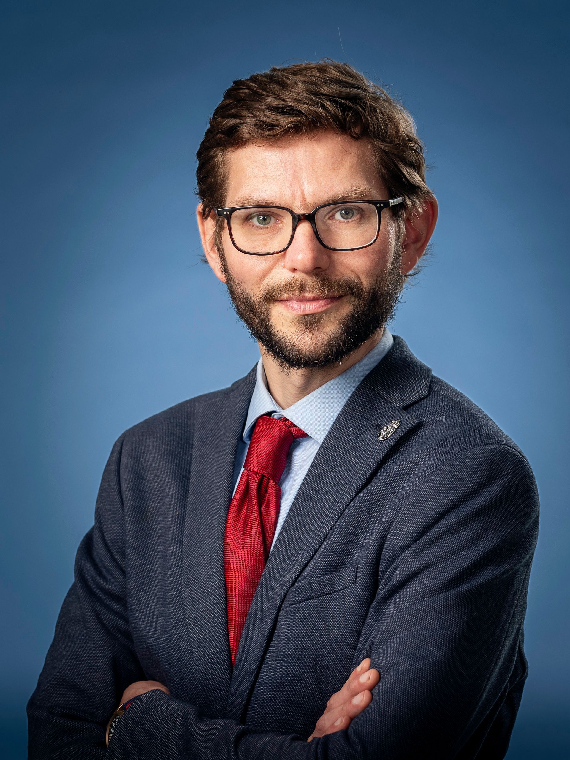 Man in pak met bril en rode stropdas, armen over elkaar, glimlachend tegen een blauwe achtergrond.