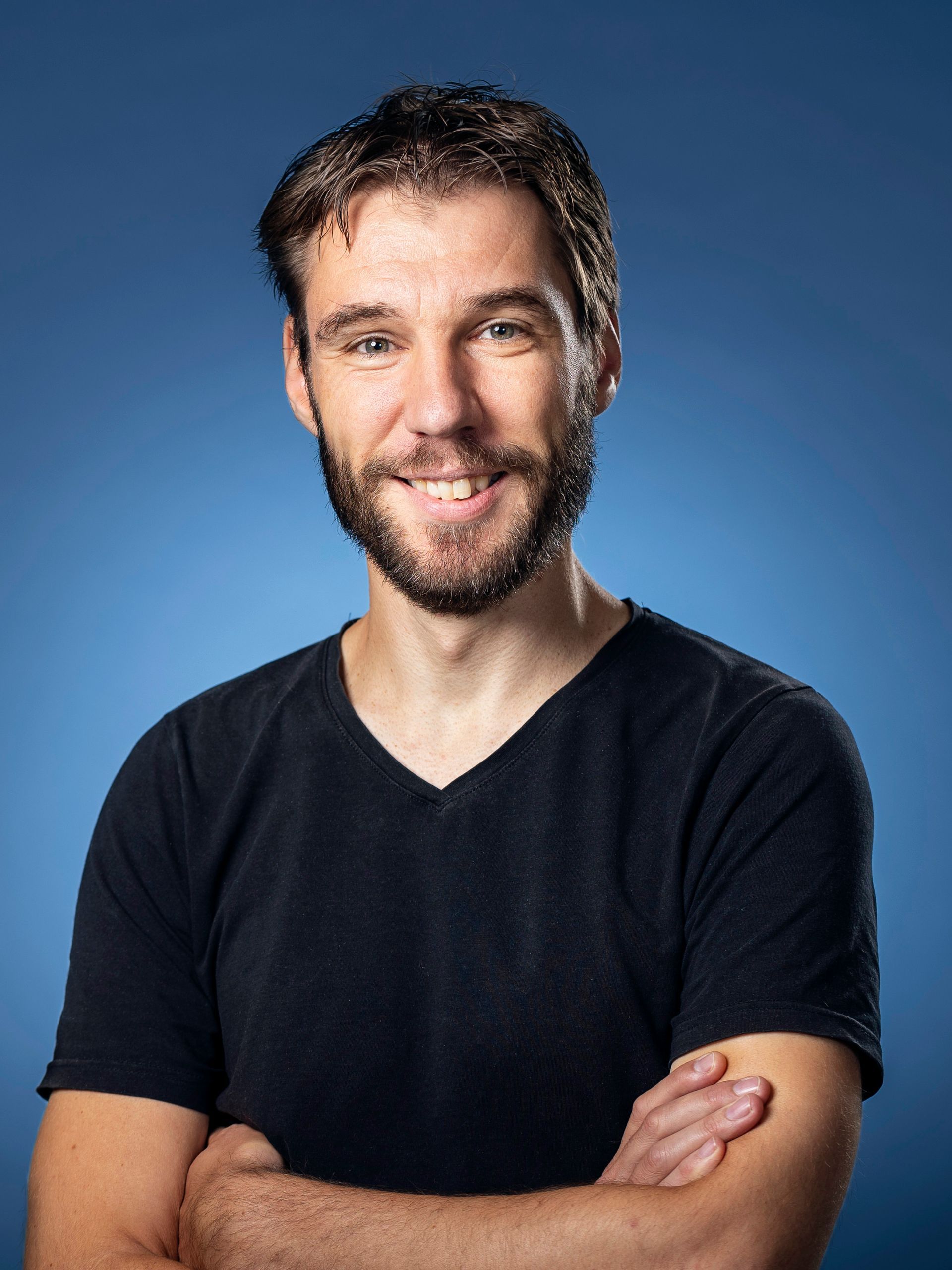 Een man met stoppelbaardje glimlacht, met de armen over elkaar, tegen een blauwe achtergrond.