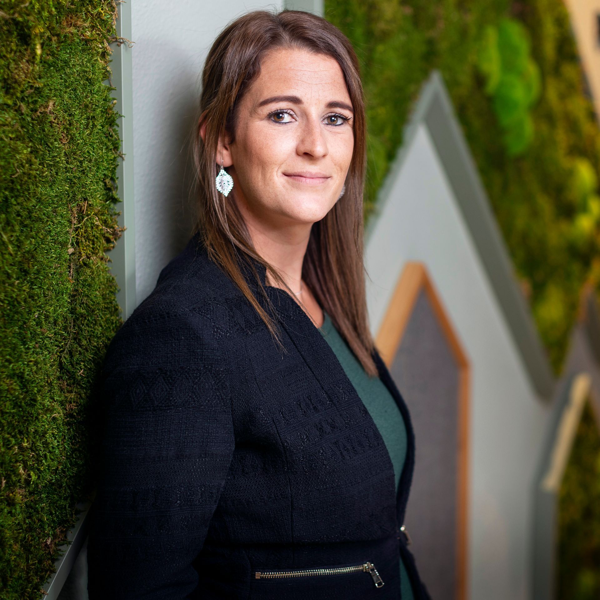 Vrouw in een blazer, glimlachend, leunend tegen een met mos begroeide muur.