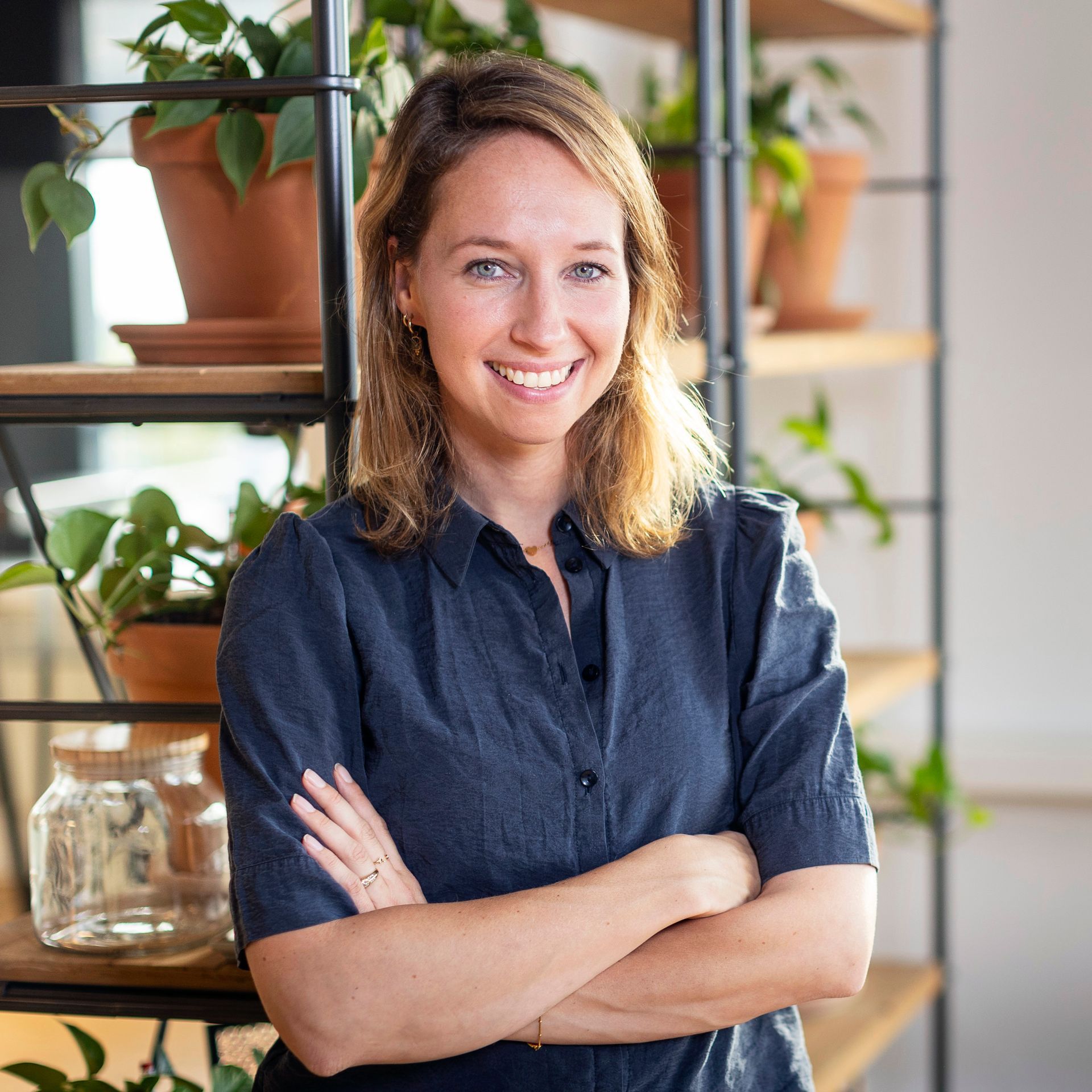 Een vrouw met gekruiste armen glimlacht terwijl ze bij een houten plank met planten staat.