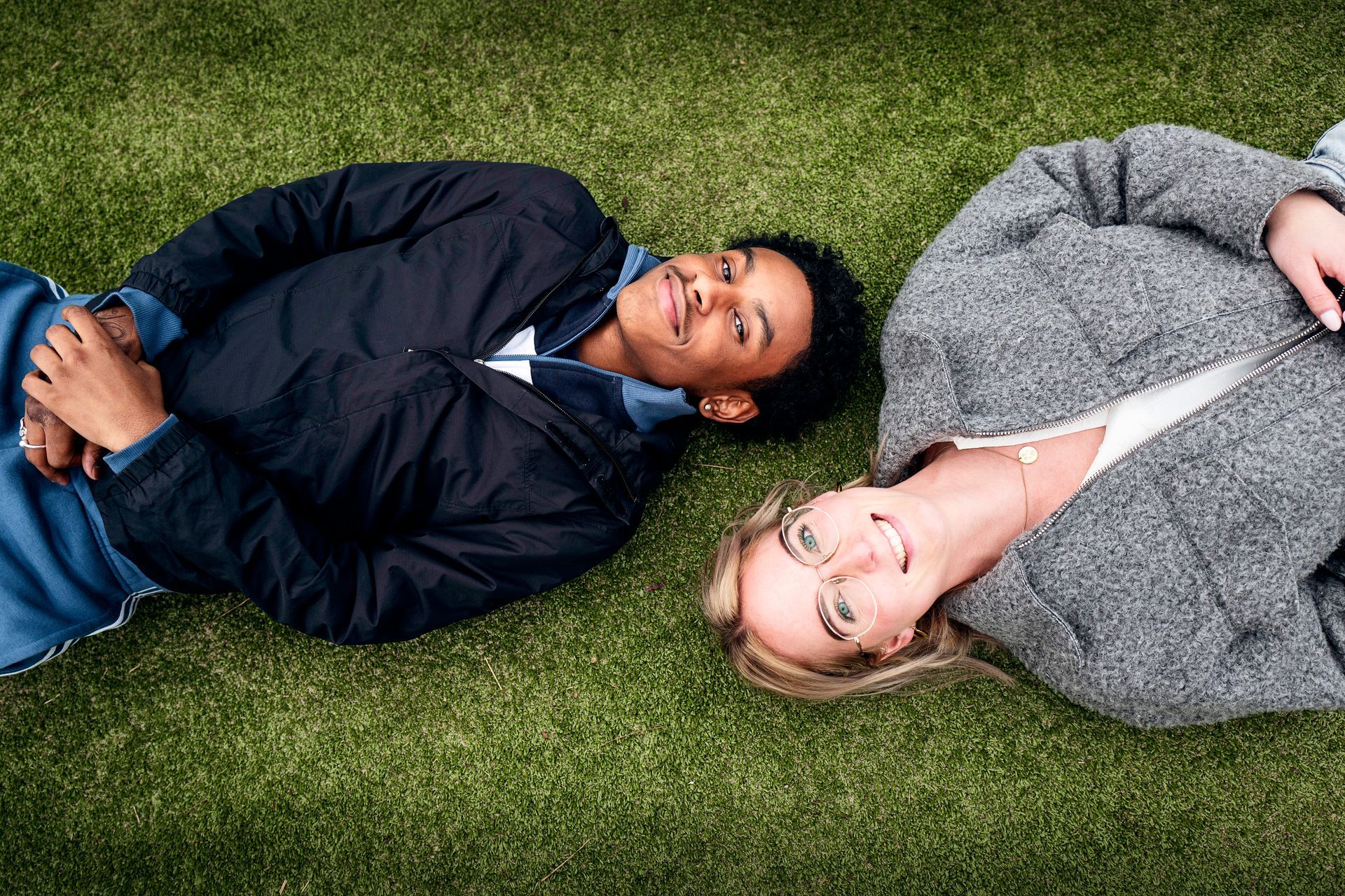 Een jonge man en een iets oudere vrouw liggen op hun rug op kunstgras en kijken omhoog. 