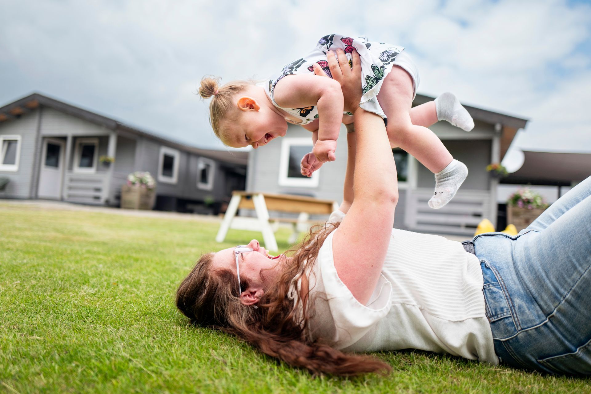 Een vrouw ligt in het gras en tilt een kind op; beiden lachen, vlakbij grijze huizen.
