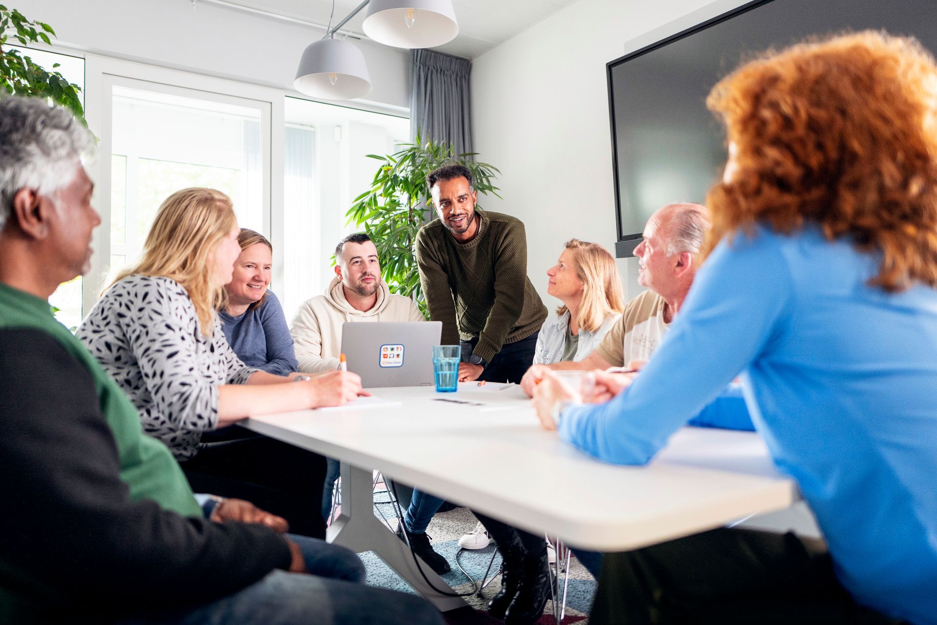 Een groep mensen zit in een vergadering rond een witte tafel en voert een discussie. 