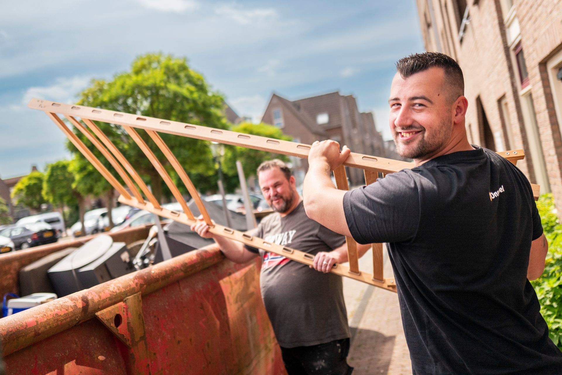 Twee mannen tillen een houten ladder uit een afvalcontainer en staan ​​lachend buiten.