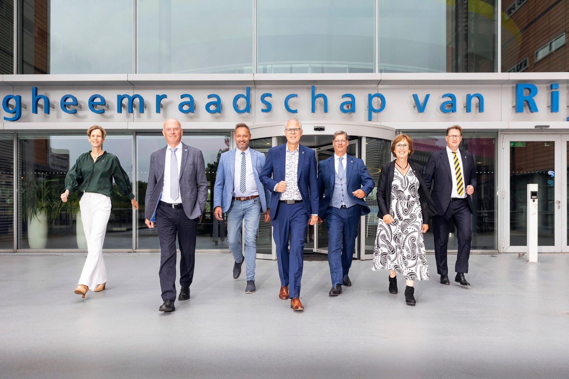 Een groep mensen in zakelijke kleding loopt richting de camera voor een gebouw met blauwe tekst.