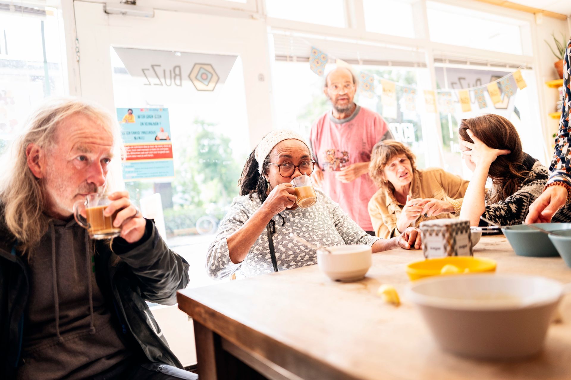Mensen zitten rond een tafel in buurthuis en drinken uit kopjes. Zonlicht stroomt naar binnen.