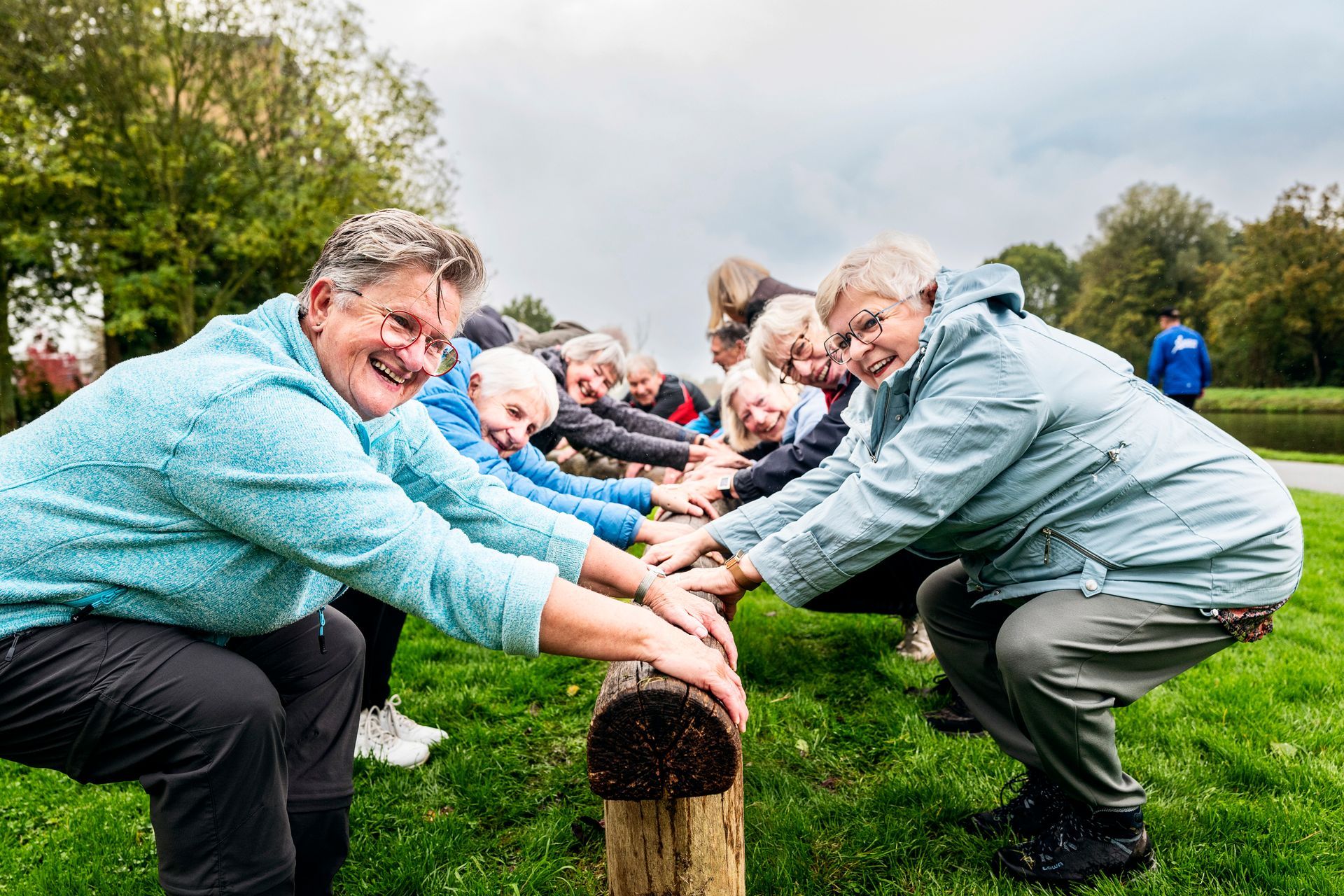 Een groep mensen in vrijetijdskleding zit gehurkt op een boomstam in een park en lacht.