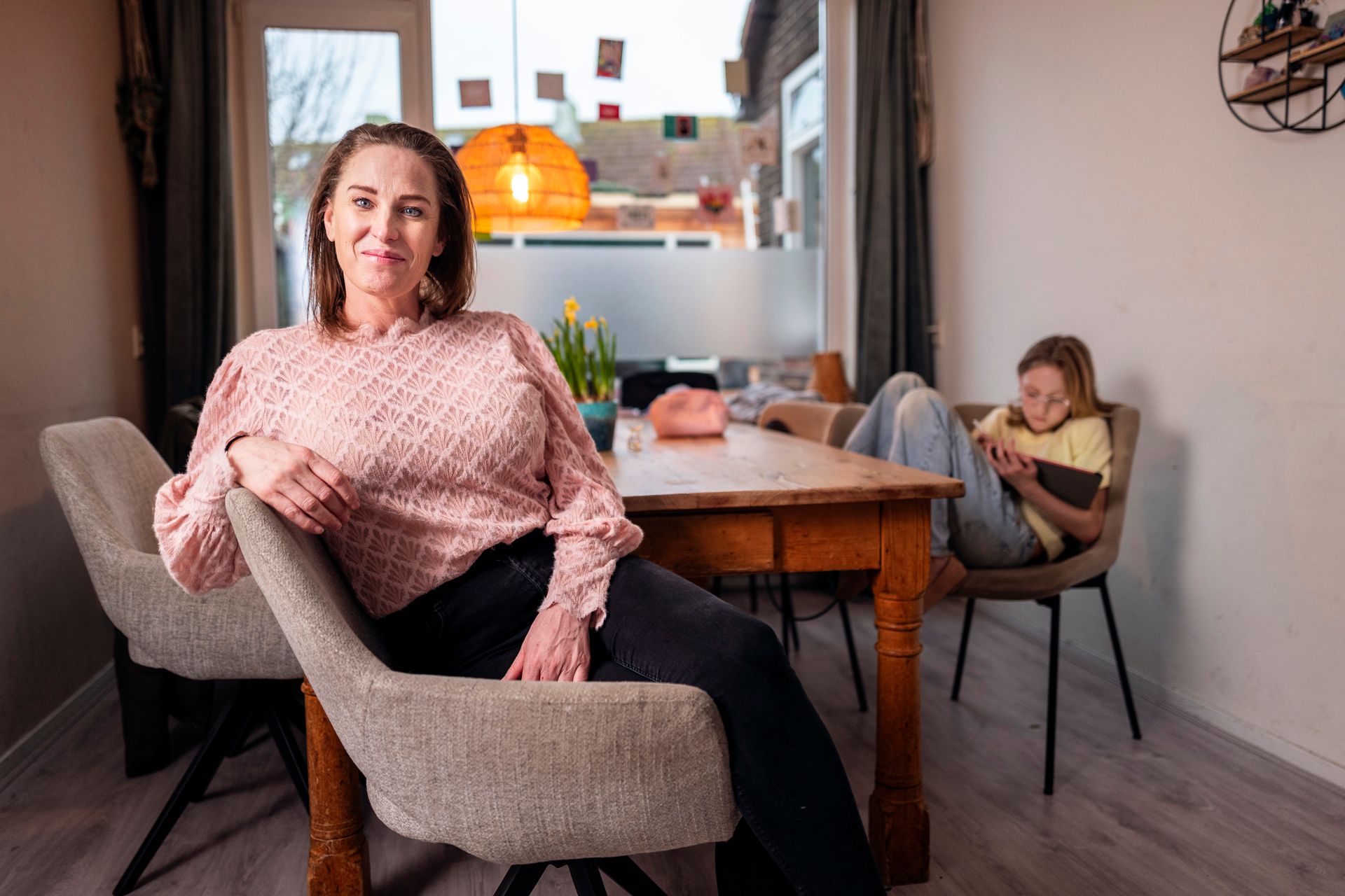 Een vrouw in een roze trui zit glimlachend voor een eettafel. Op de achtergrondeen tiener.