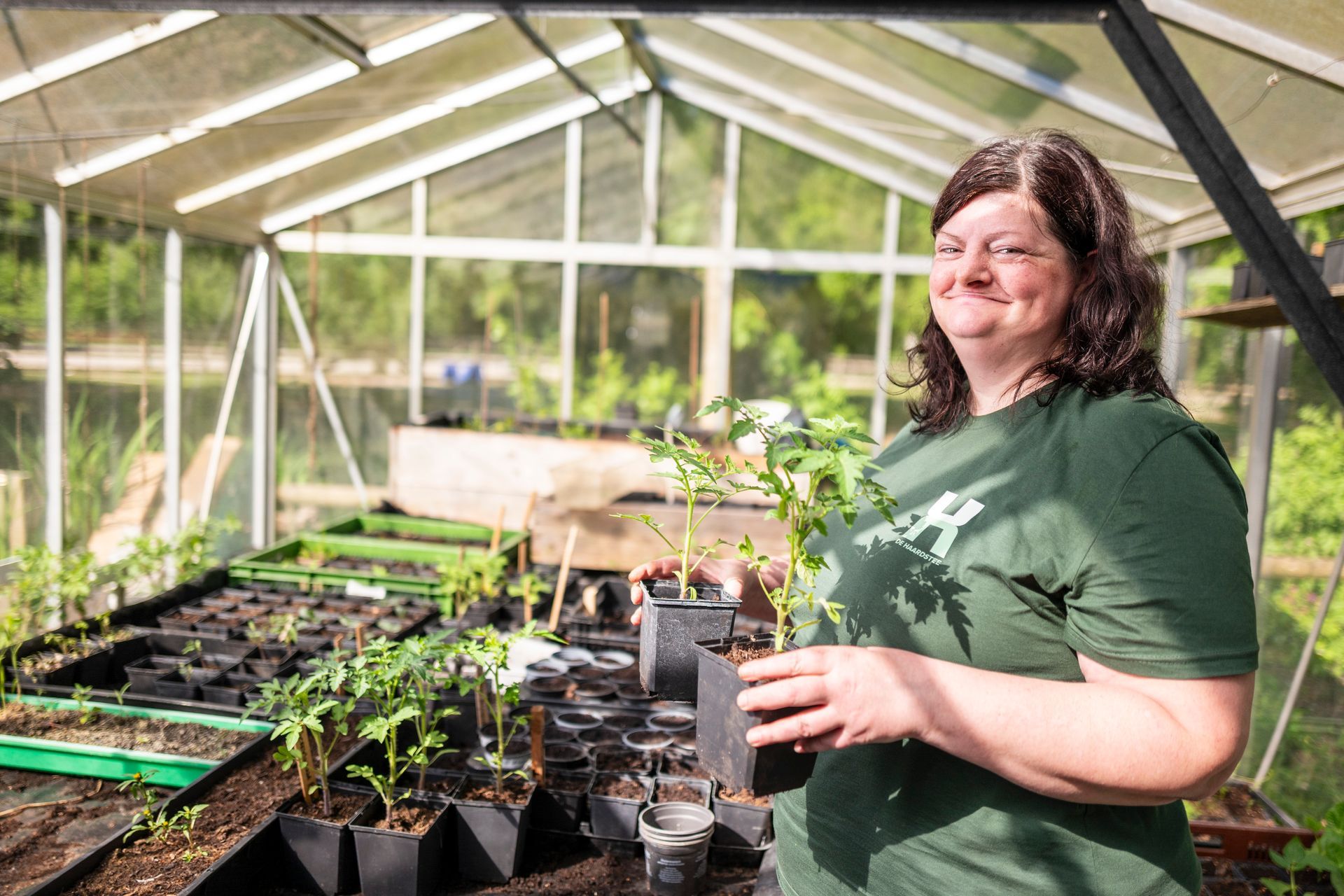 Een vrouw in een groene polo lacht in een kas en houdt een potplant vast. Zaailingen zijn zichtbaar.