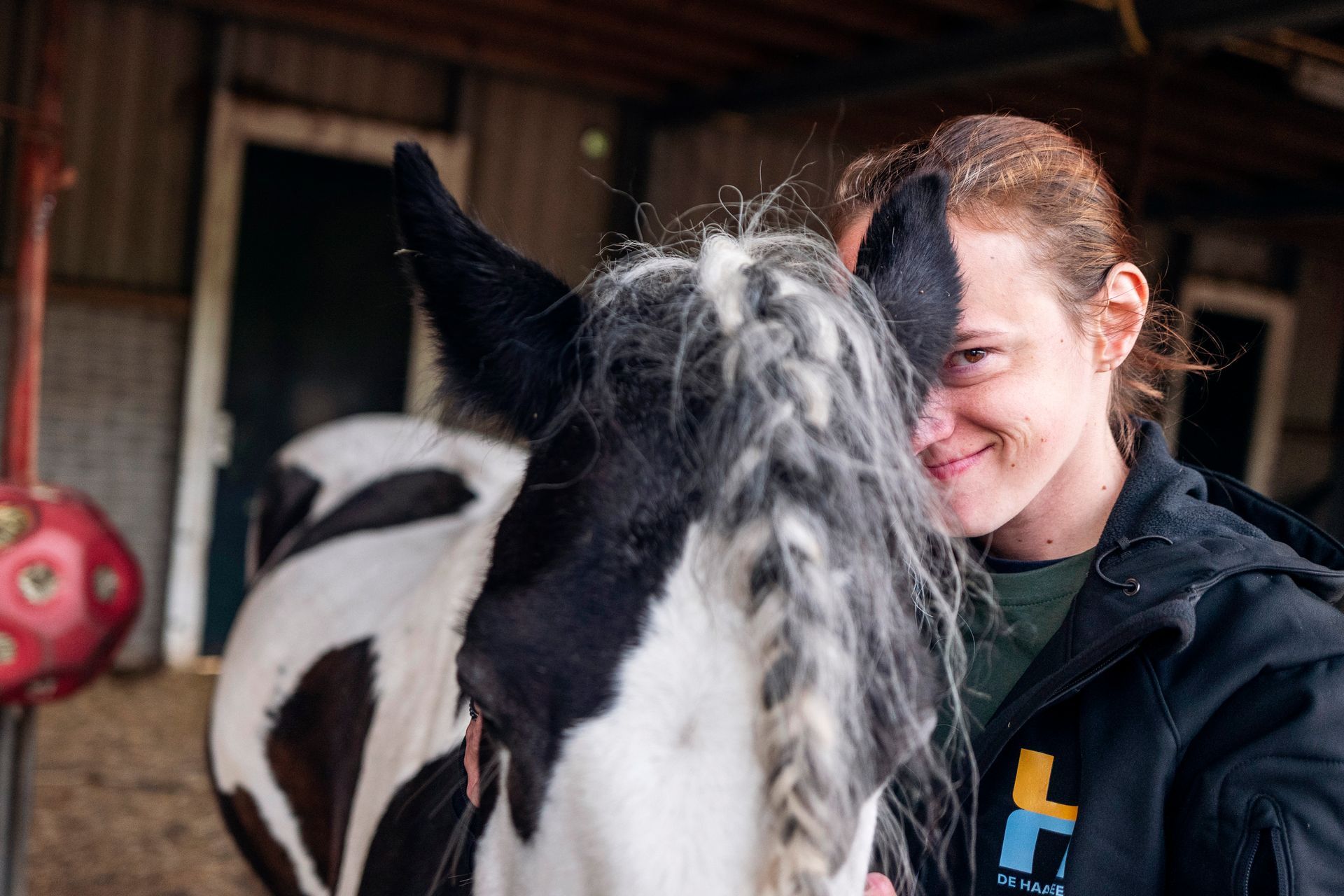 Een lachende vrouw staat naast een zwart-wit paard in een schuur.