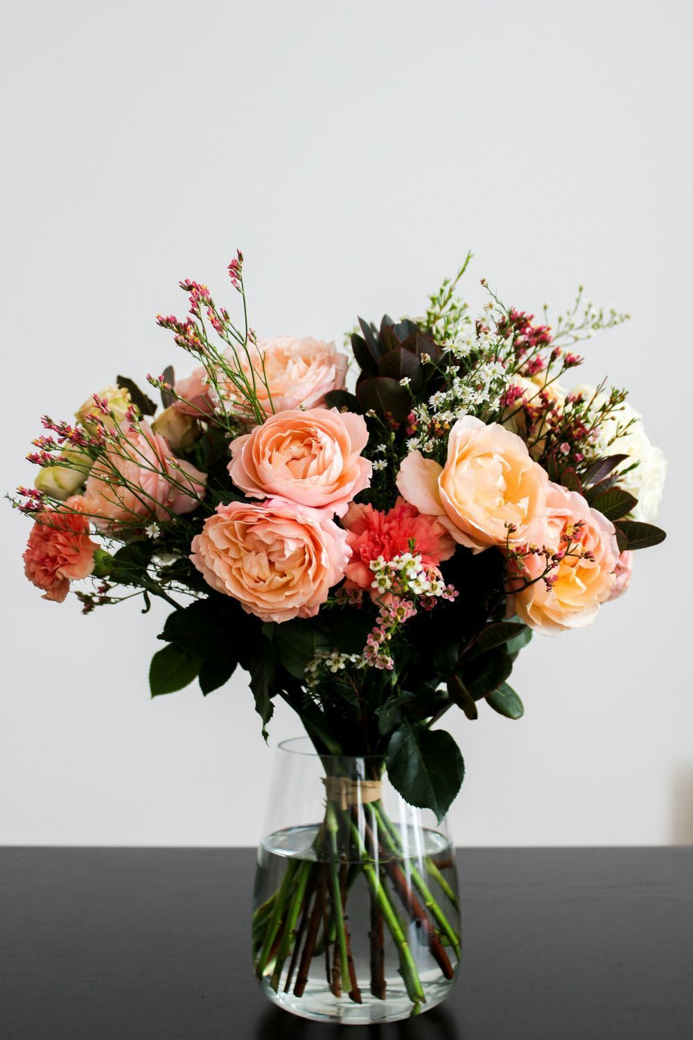 Bouquet of peach and pink flowers in a clear glass vase on a dark table against a white background