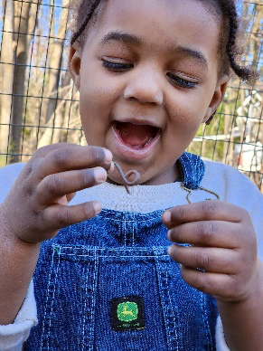 Child smiles, holding a small worm outdoors. Child wears denim overalls.