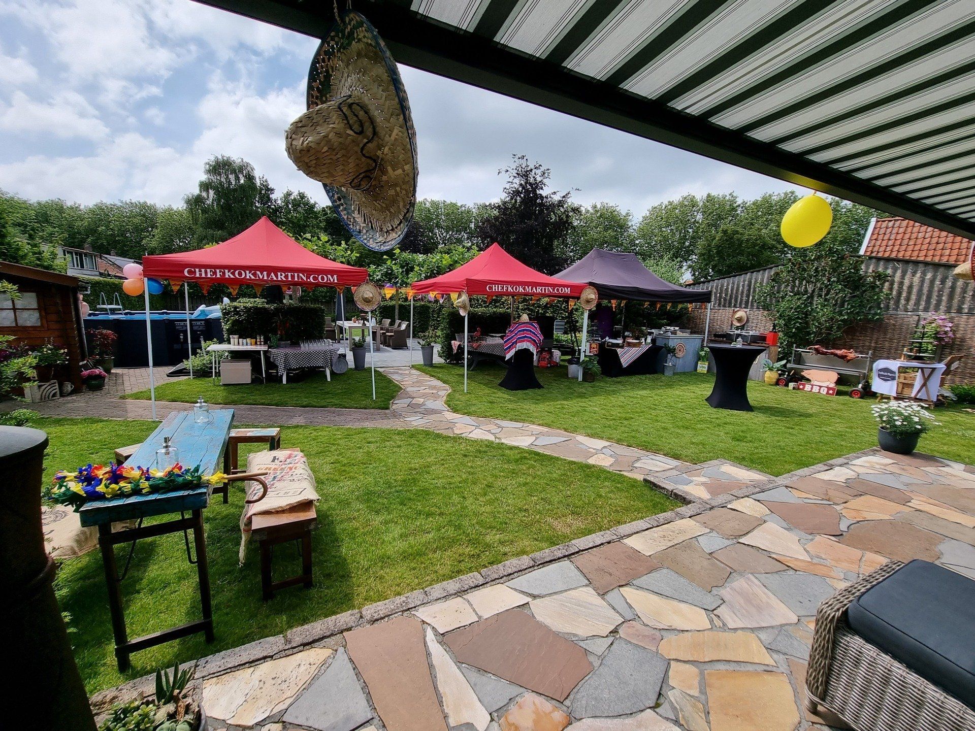 Tuinfeest met rode partytenten, tafels, ballonnen en een stenen terras.