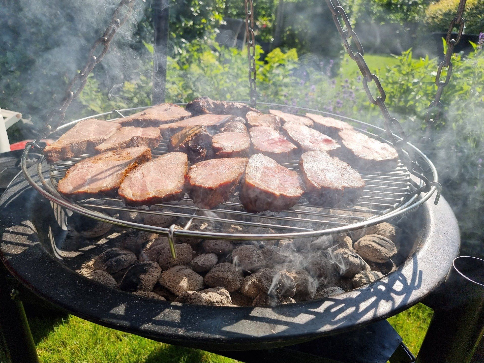 Vlees wordt gegrild boven hete kolen op een buitenbarbecue, waarbij rook opstijgt.
