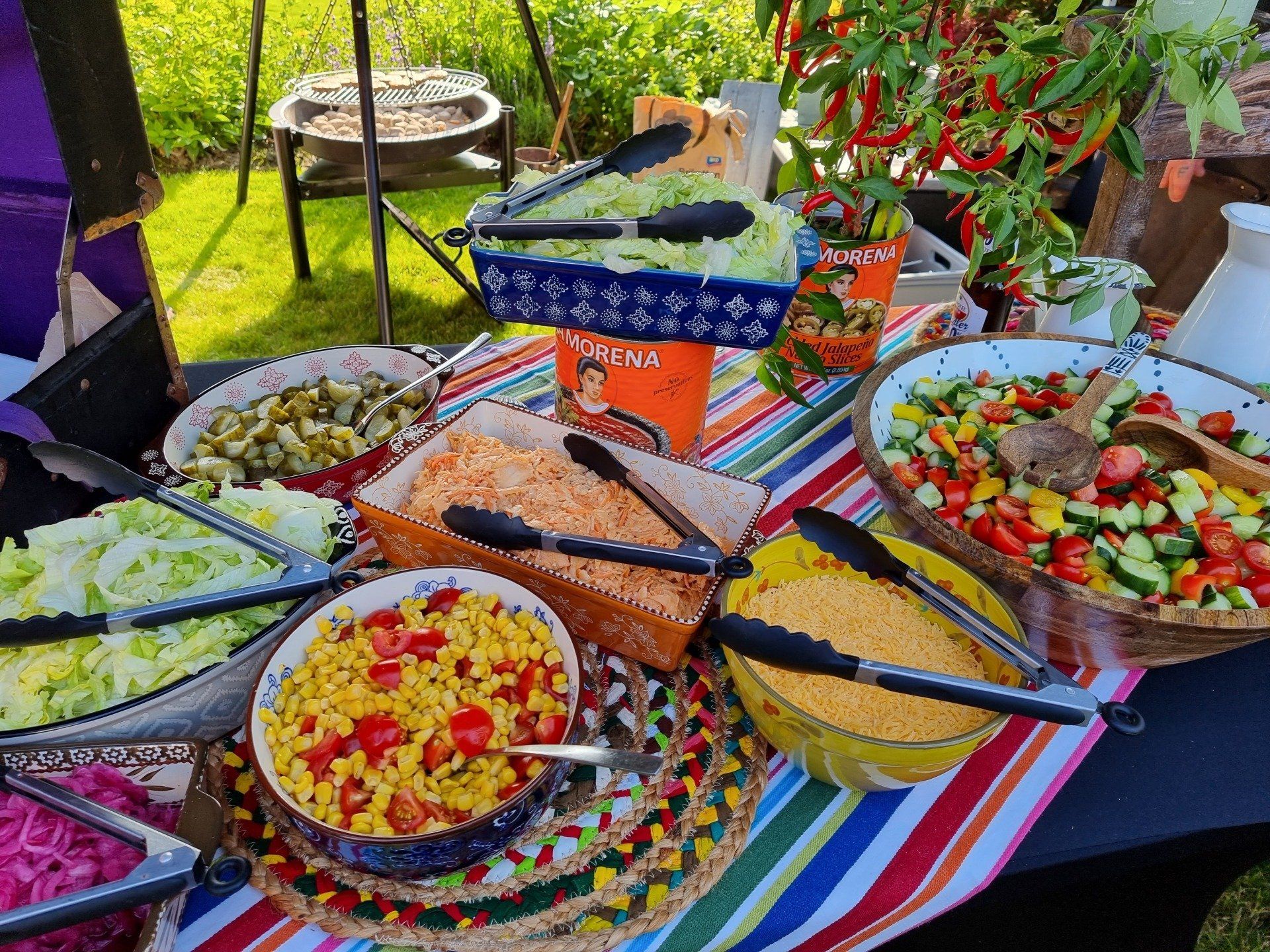 Een kleurrijk buffet uitgestald op een tafel buiten; diverse kleurrijke saladeschalen en een grill op de achtergrond.