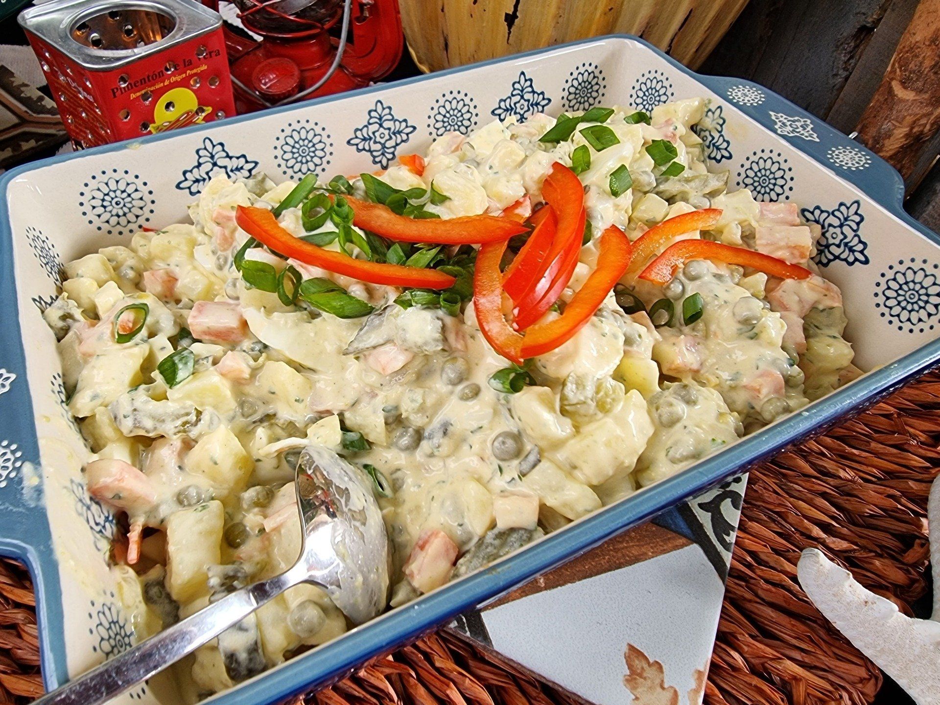 Aardappelsalade in een decoratieve blauwe schaal, gegarneerd met rode paprika en lente-uitjes.