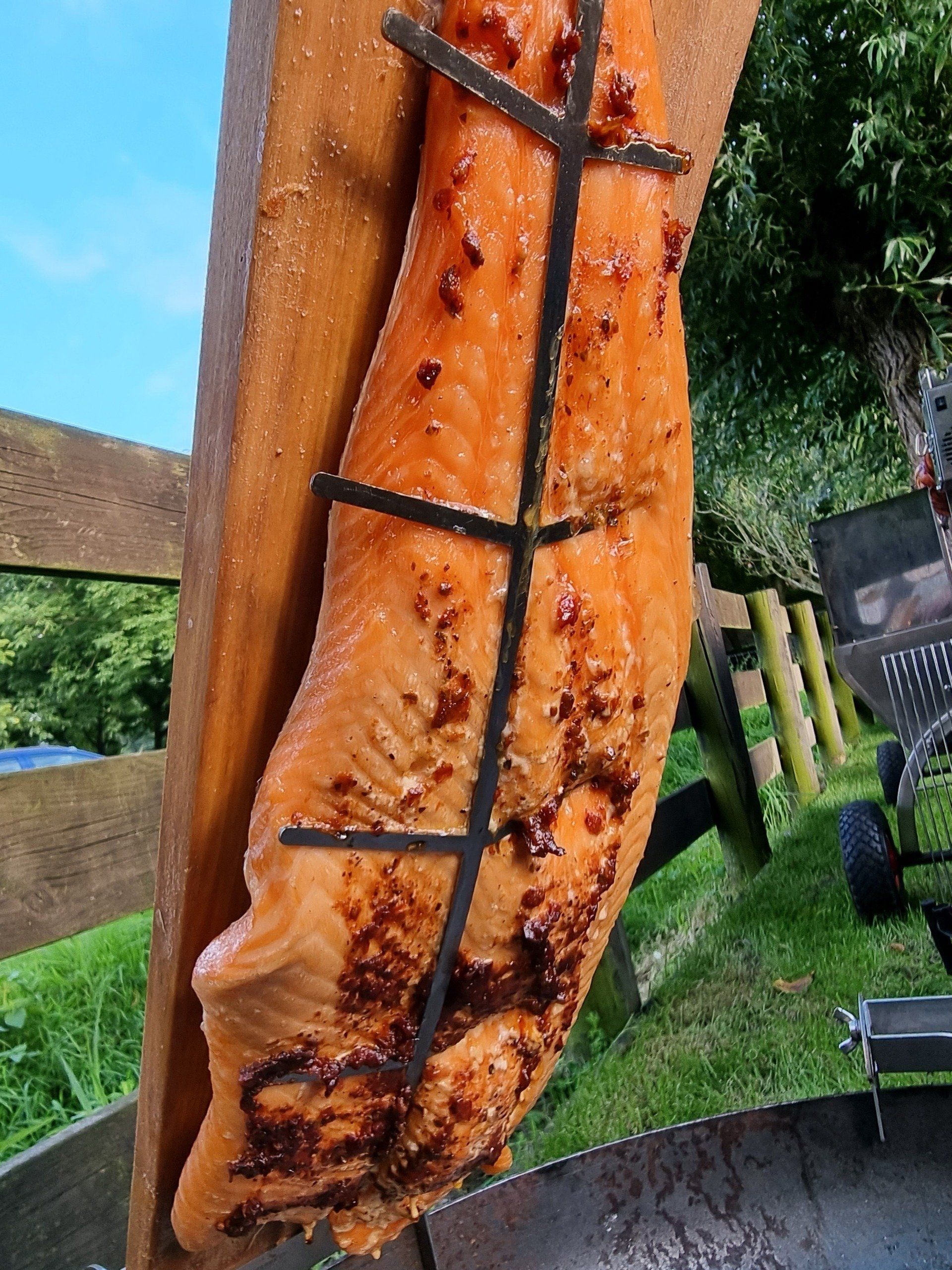 Gerookte zalm op een houten plank, bevestigd aan een metalen frame, buiten, met groen en een hek op de achtergrond.