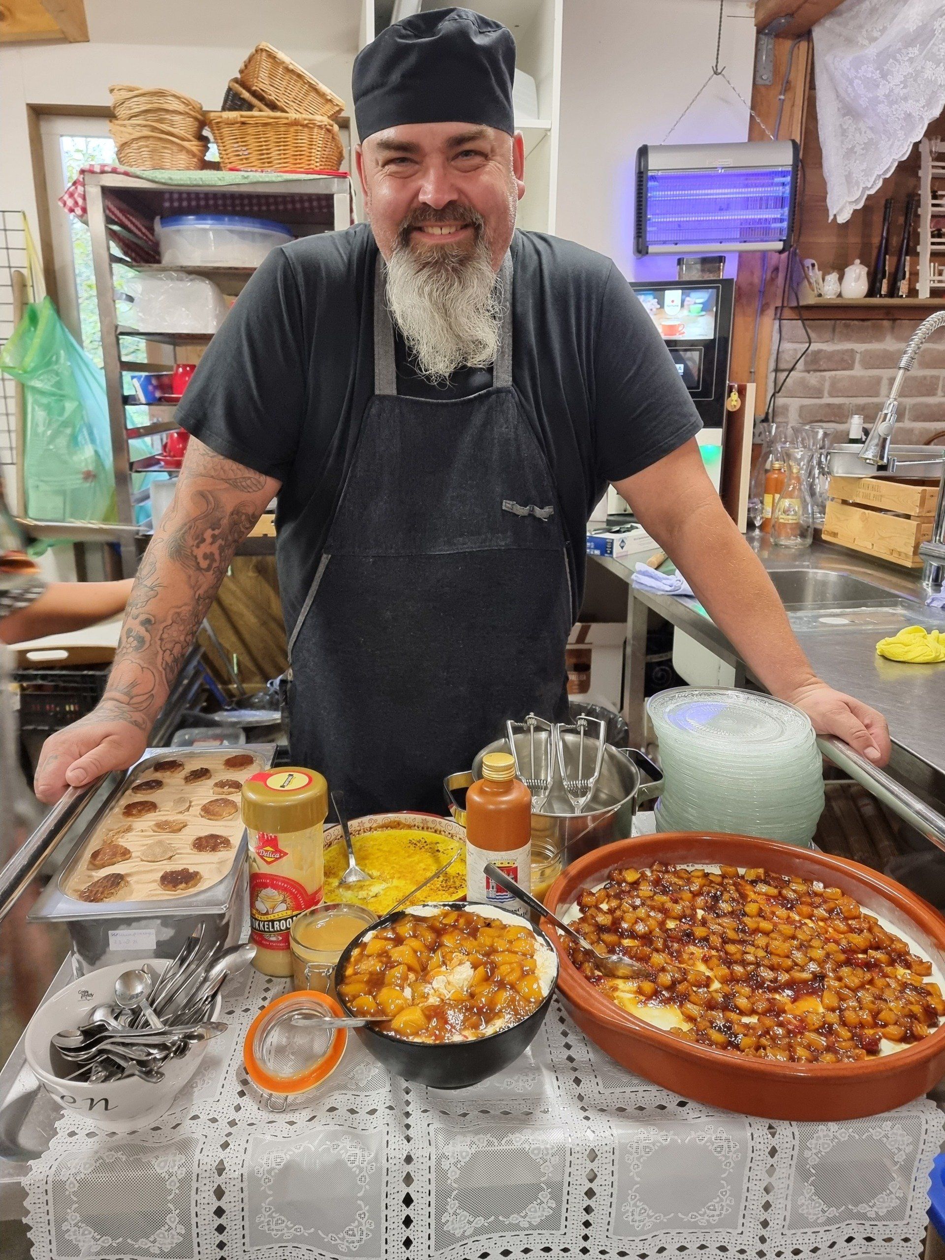 Chefkok Martin staat achter een tafel vol eten in een keuken. Hij glimlacht en draagt ​​een zwart schort en een koksmuts.