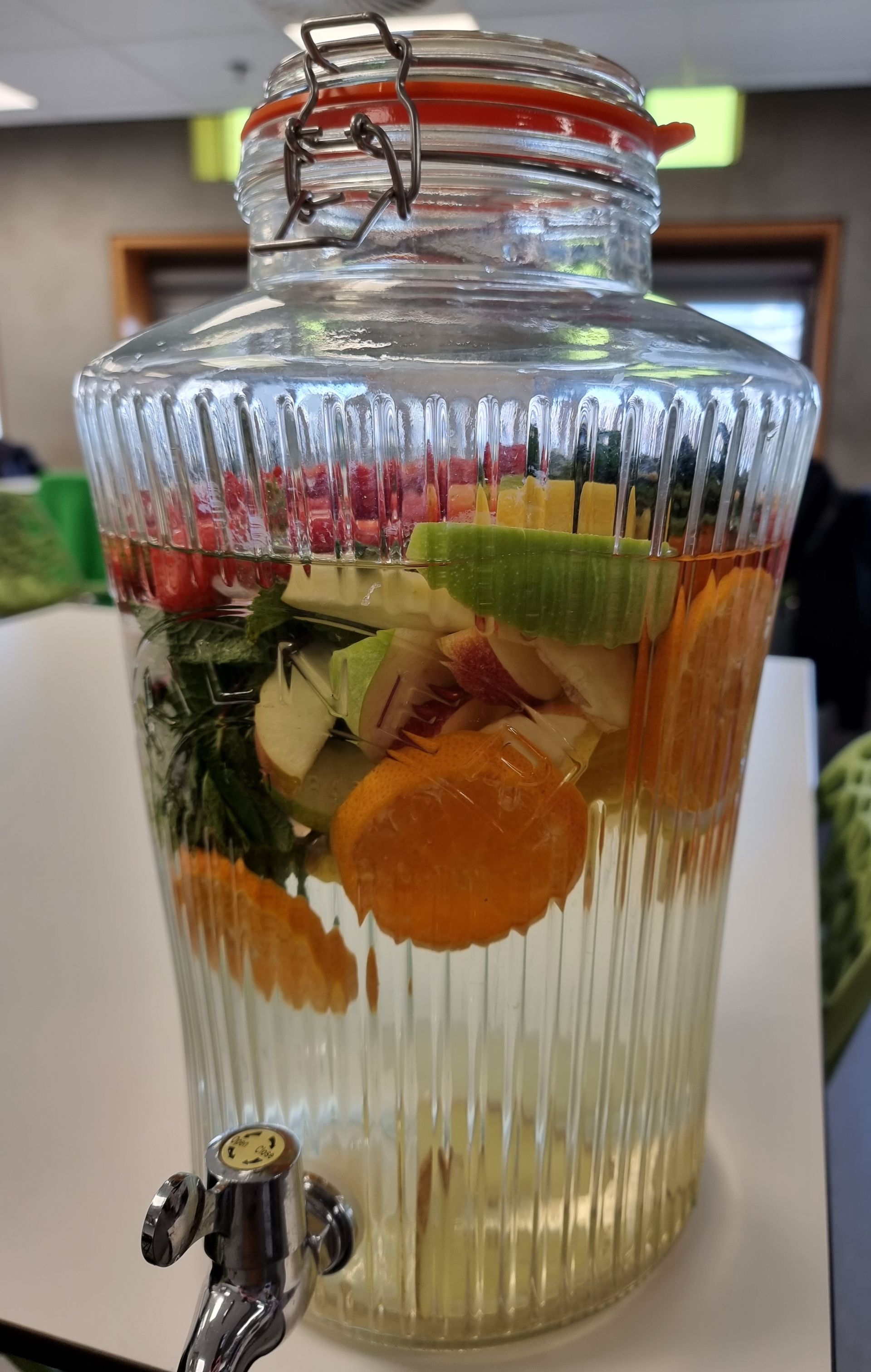 Glazen drankdispenser gevuld met fruit en water. Sinaasappel, appel en kruiden zijn zichtbaar.