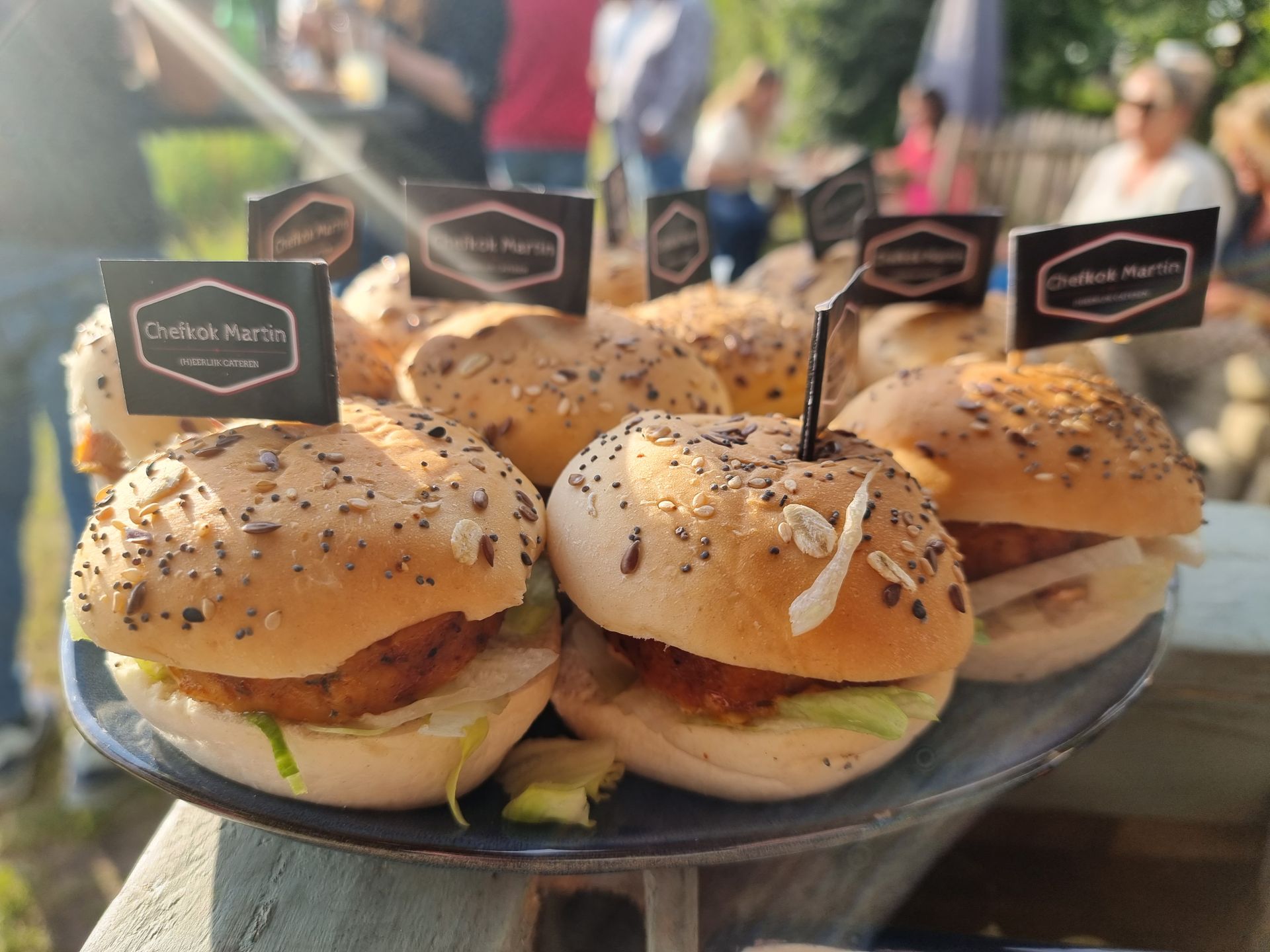 Mini broodjes huisgemaakte grillworst op een schaal, elk met een prikker met logo, buiten op een evenement.