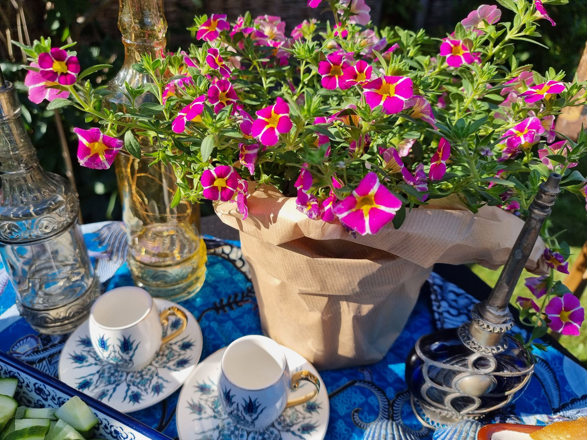 Roze bloemen in een papieren pot op een blauwe tafel, met koffiekopjes, flessen en een vijzel en stamper.