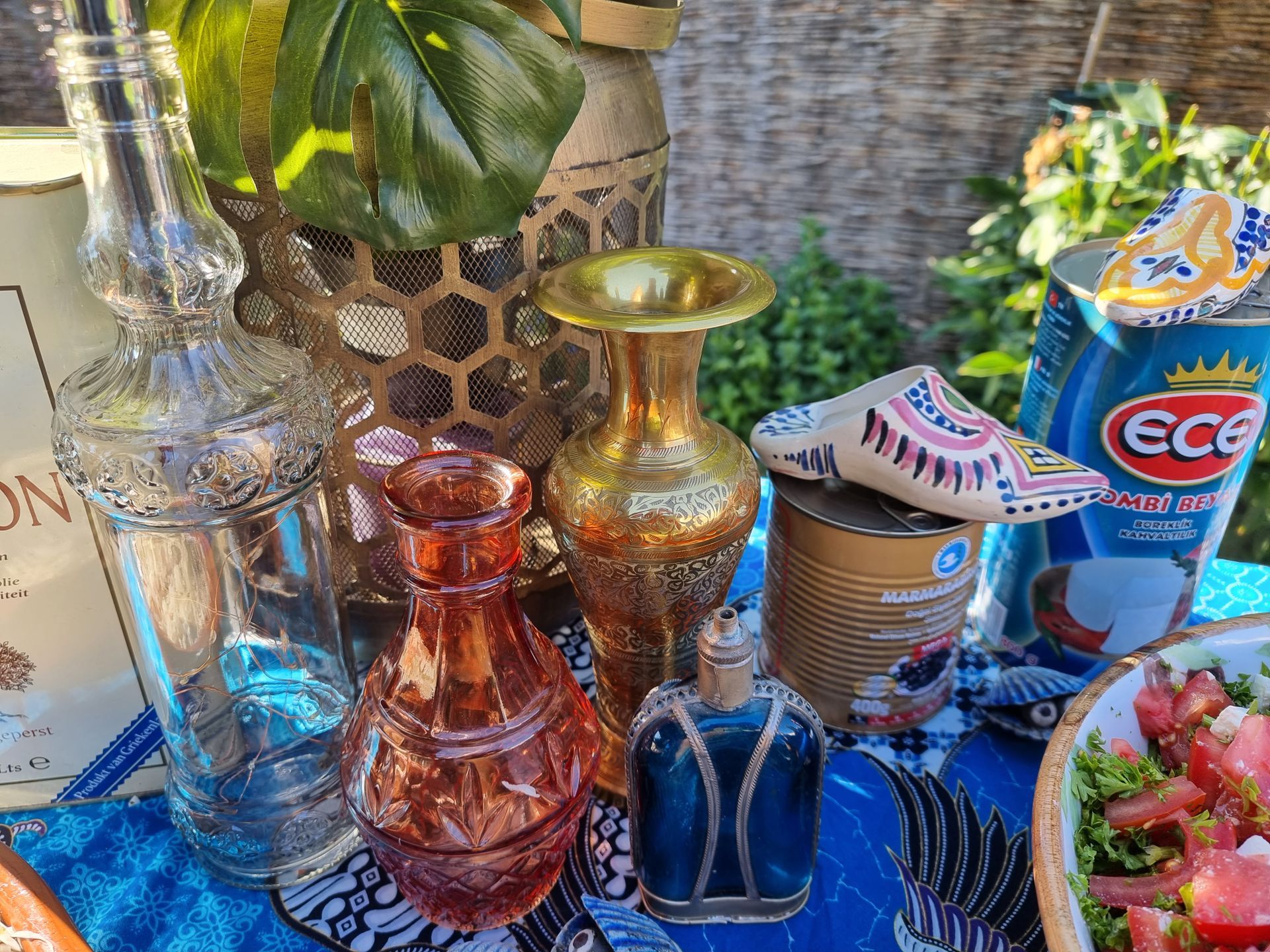 Diverse glazen flessen en vazen ​​op een blauw tafelkleed met patroon, met eten en planten op de achtergrond.