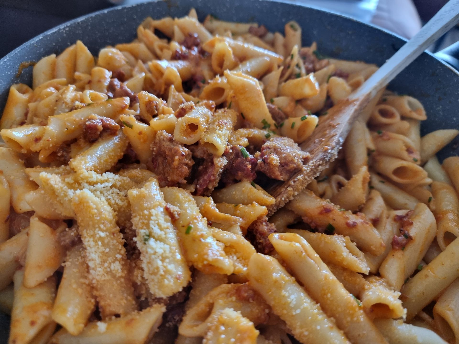 Penne pasta met rode saus, worst en Parmezaanse kaas in een pan, geroerd met een houten lepel.
