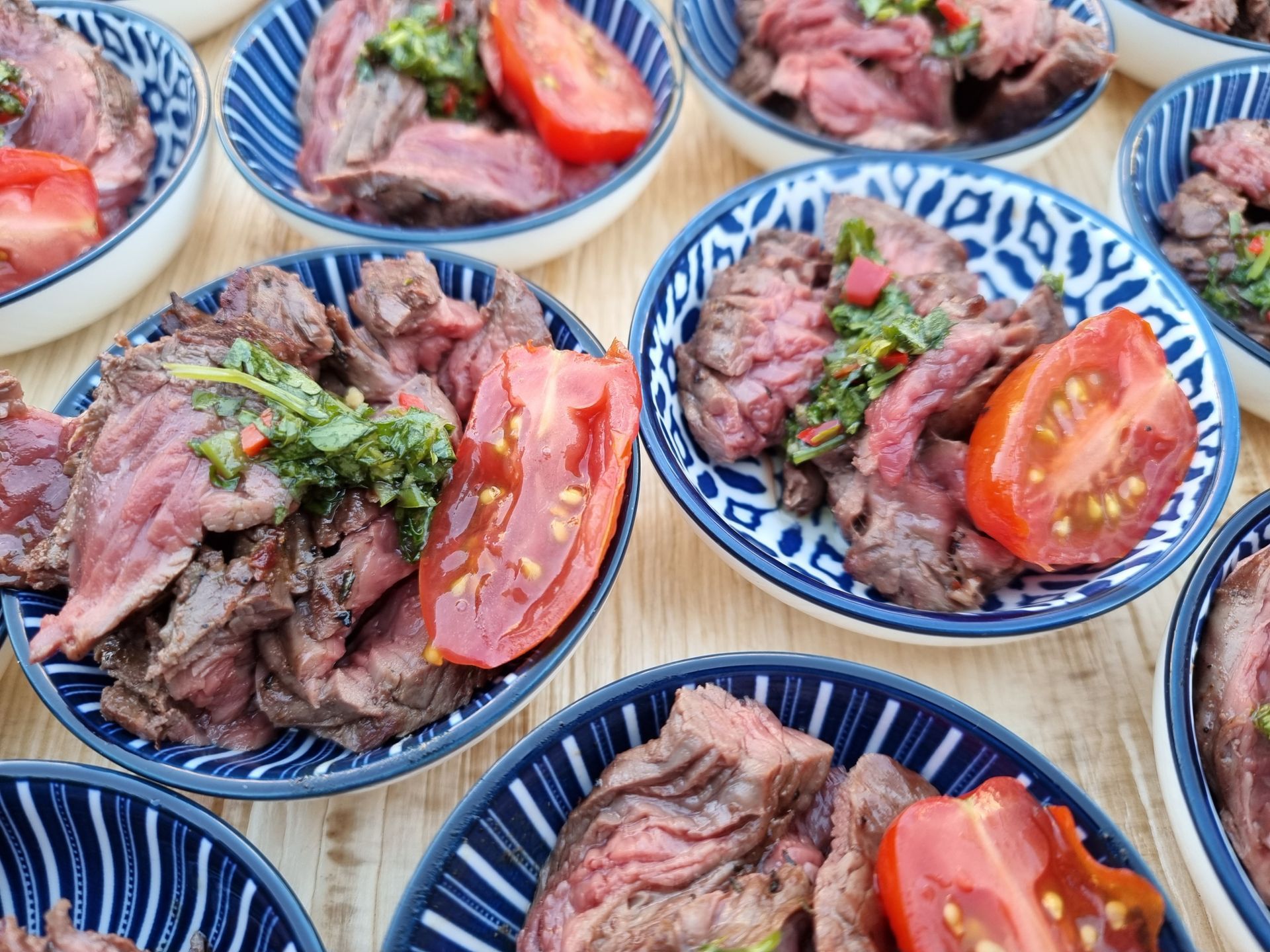 Schalen met gesneden bavette, tomaat en groene saus, gerangschikt op een houten ondergrond.