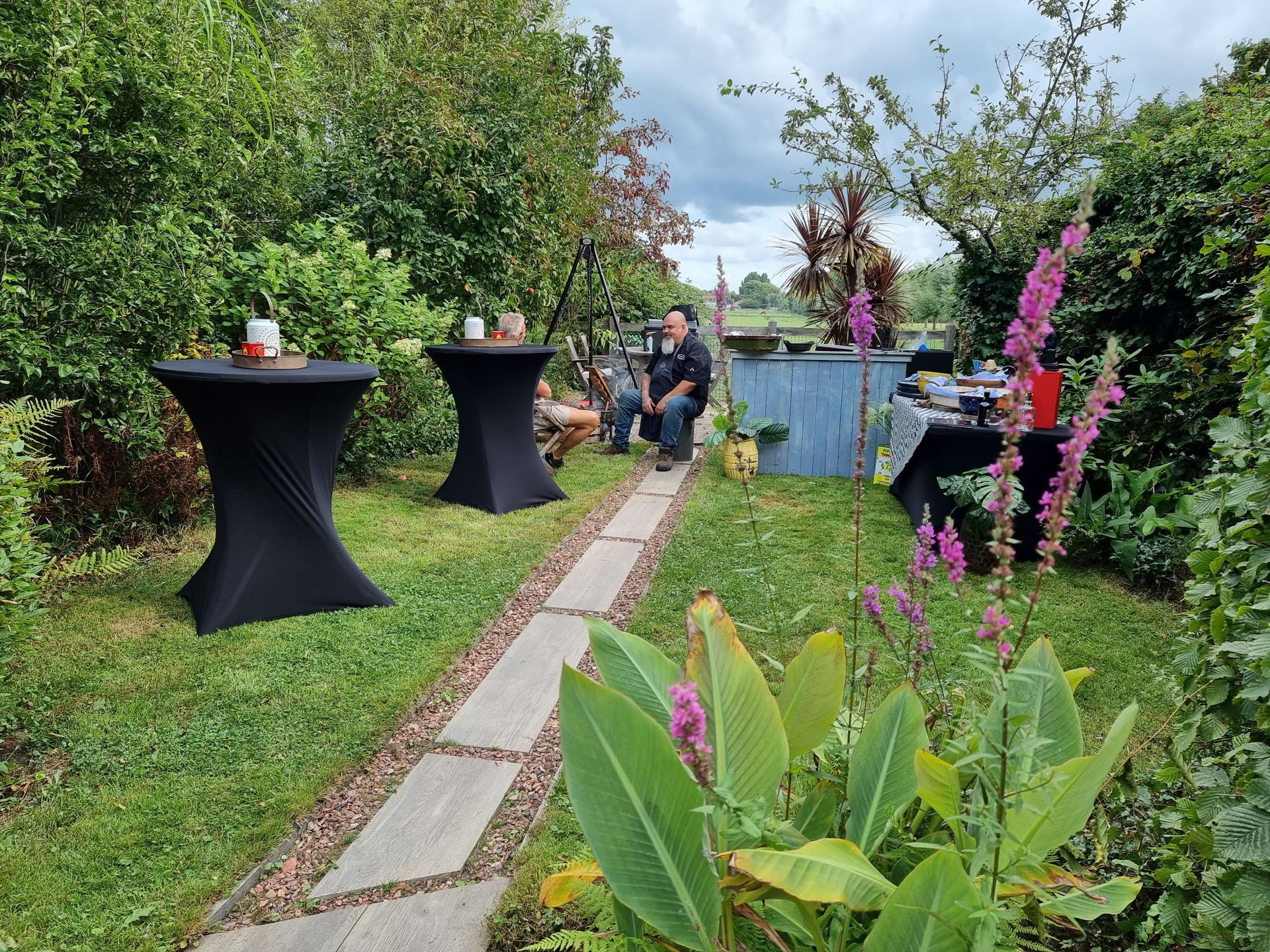 Buitenevenement: pad door de tuin met kleine tafels, mensen en een bar op de achtergrond.