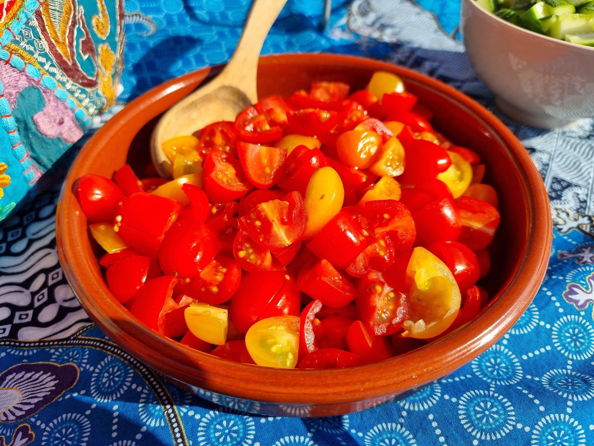 Een kom met in blokjes gesneden rode en gele tomaten, geserveerd met een houten lepel, staat op een blauw tafelkleed.