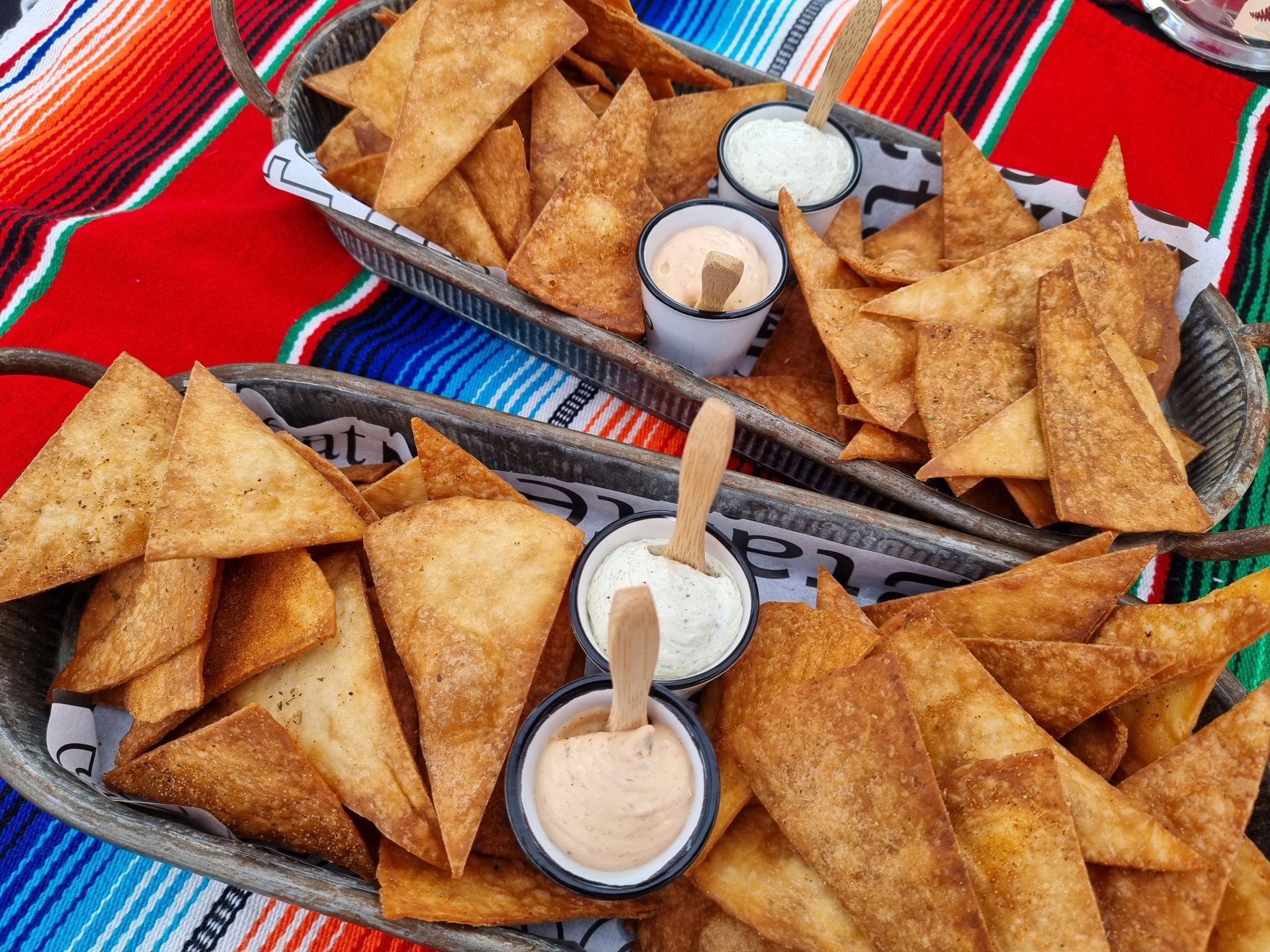 Huisgemaakte tortillachips met dipsauzen op serveerschalen, op een kleurrijk, gedessineerd tafelkleed.