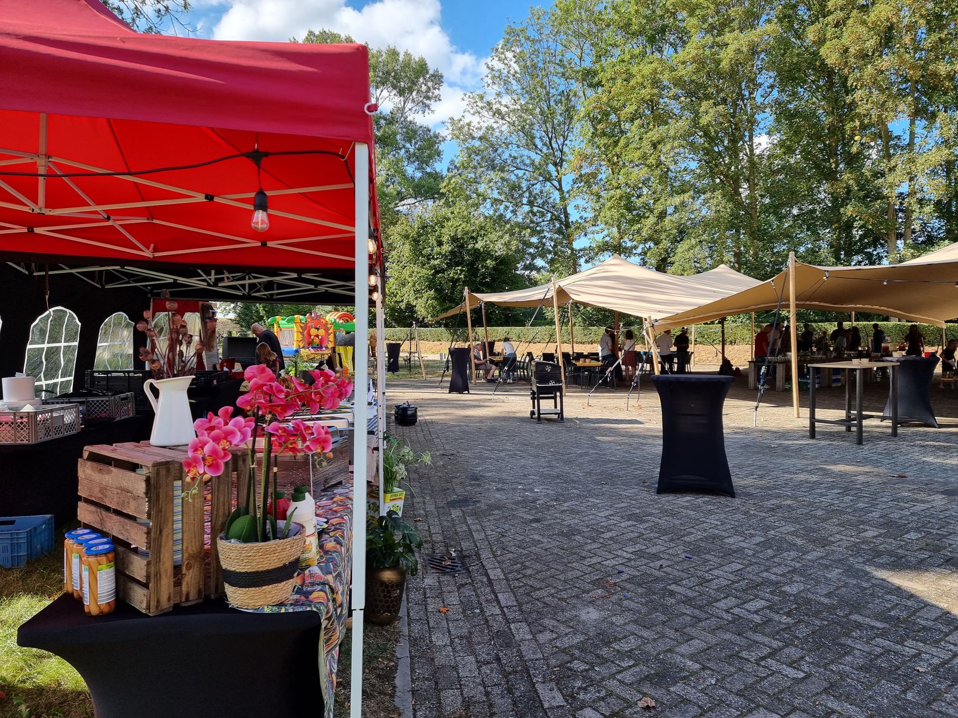 Buitenevenement met rode tent en tafels, mensen onder stretchtent, bomen op de achtergrond.
